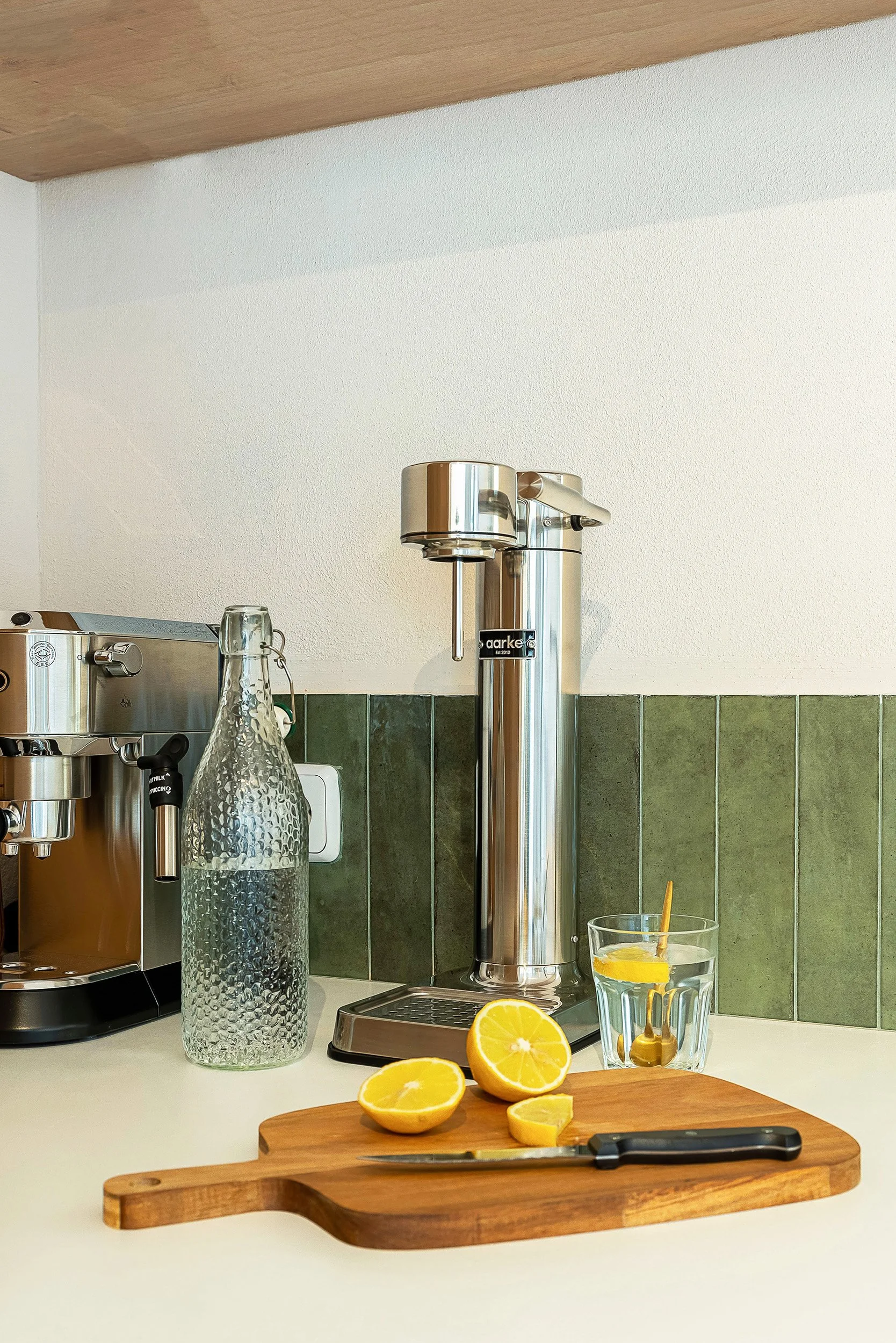 A kitchen countertop with a soda maker, an espresso machine, and a glass water bottle. A cutting board with sliced lemons and a knife is in the foreground, while a glass of water with lemon and a golden spoon is in the background.