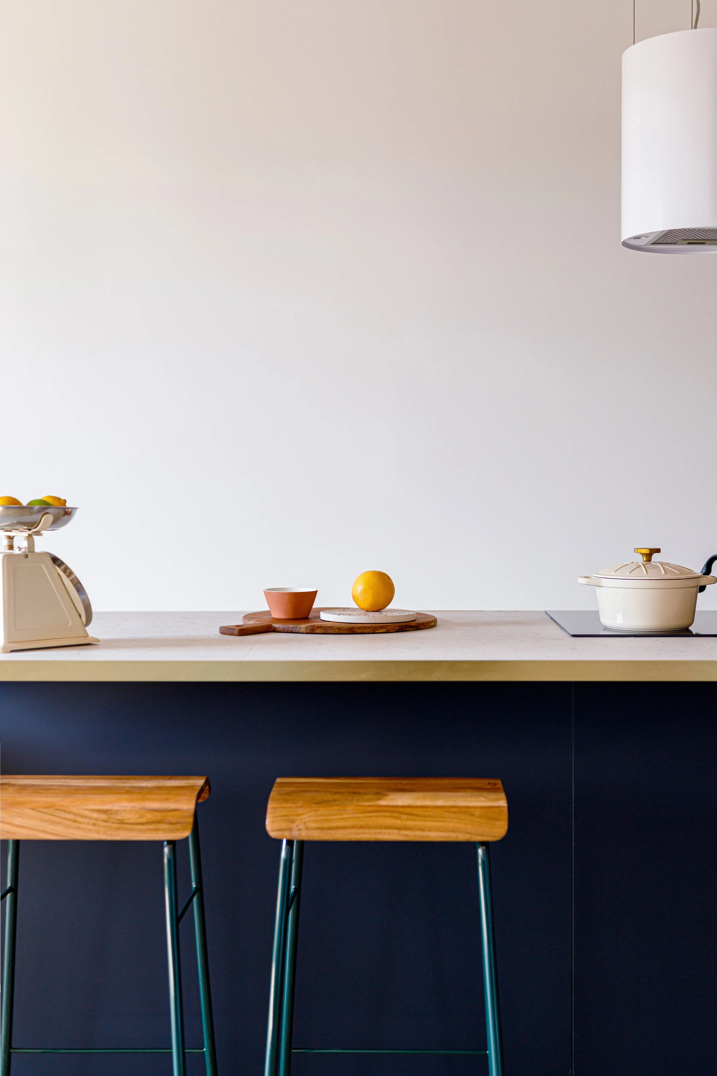 Minimalist kitchen counter with stools, a bowl of fruit on a vintage scale, a wooden cutting board with a cup and an orange, and a pot on a stove. Modern design with neutral colors.