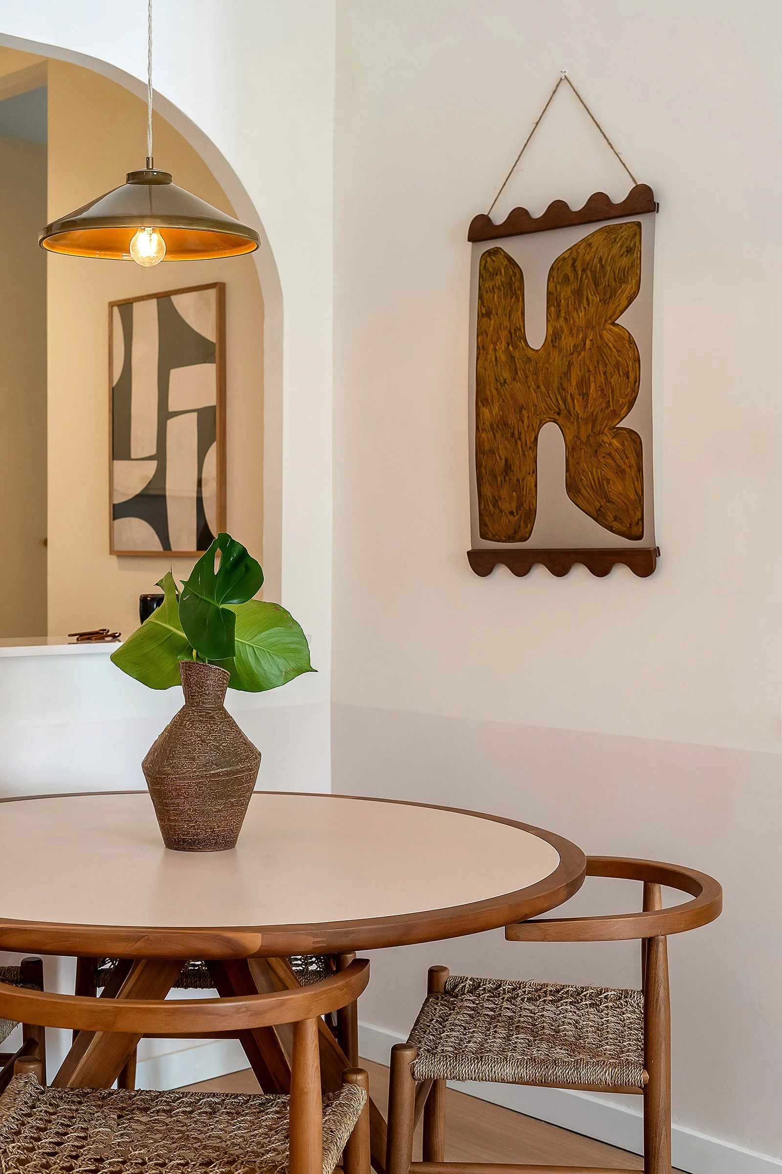Modern dining room with round wooden table, woven chairs, vase with green leaves, hanging light fixture, and abstract wall art.