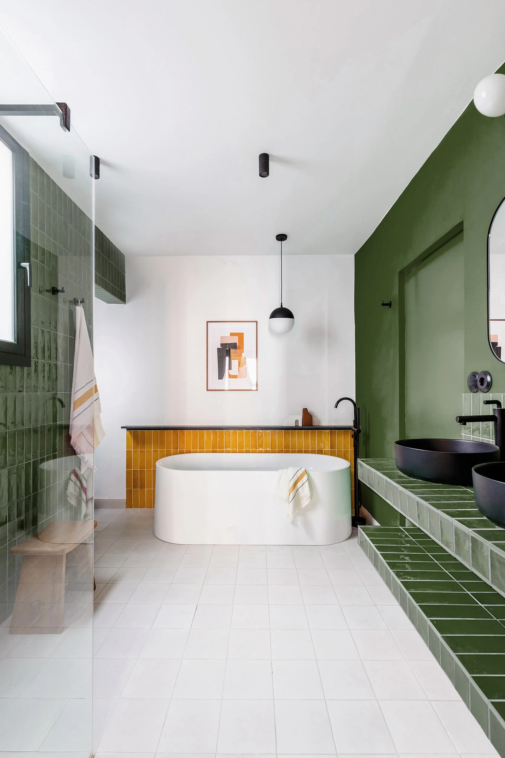 Modern bathroom with green and mustard tiles, featuring a freestanding bathtub, black sinks, a wooden bench, wall art, and a pendant light.