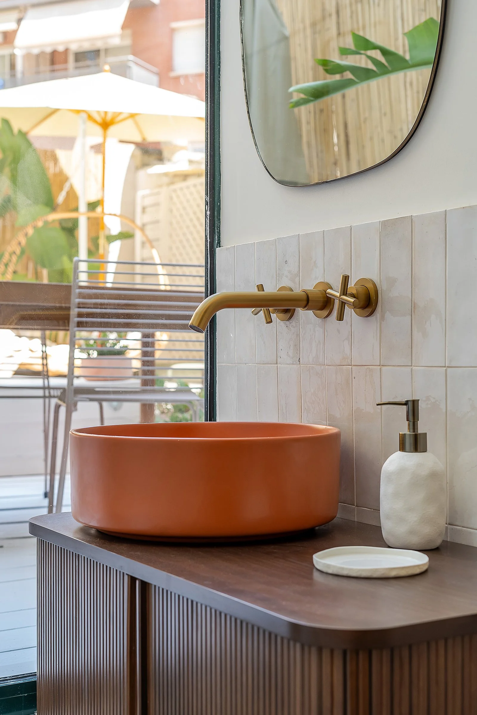 Stylish bathroom with orange vessel sink, brass wall-mounted faucet, and soap dispenser on wooden vanity. Outdoor patio visible through window with plants and furniture.