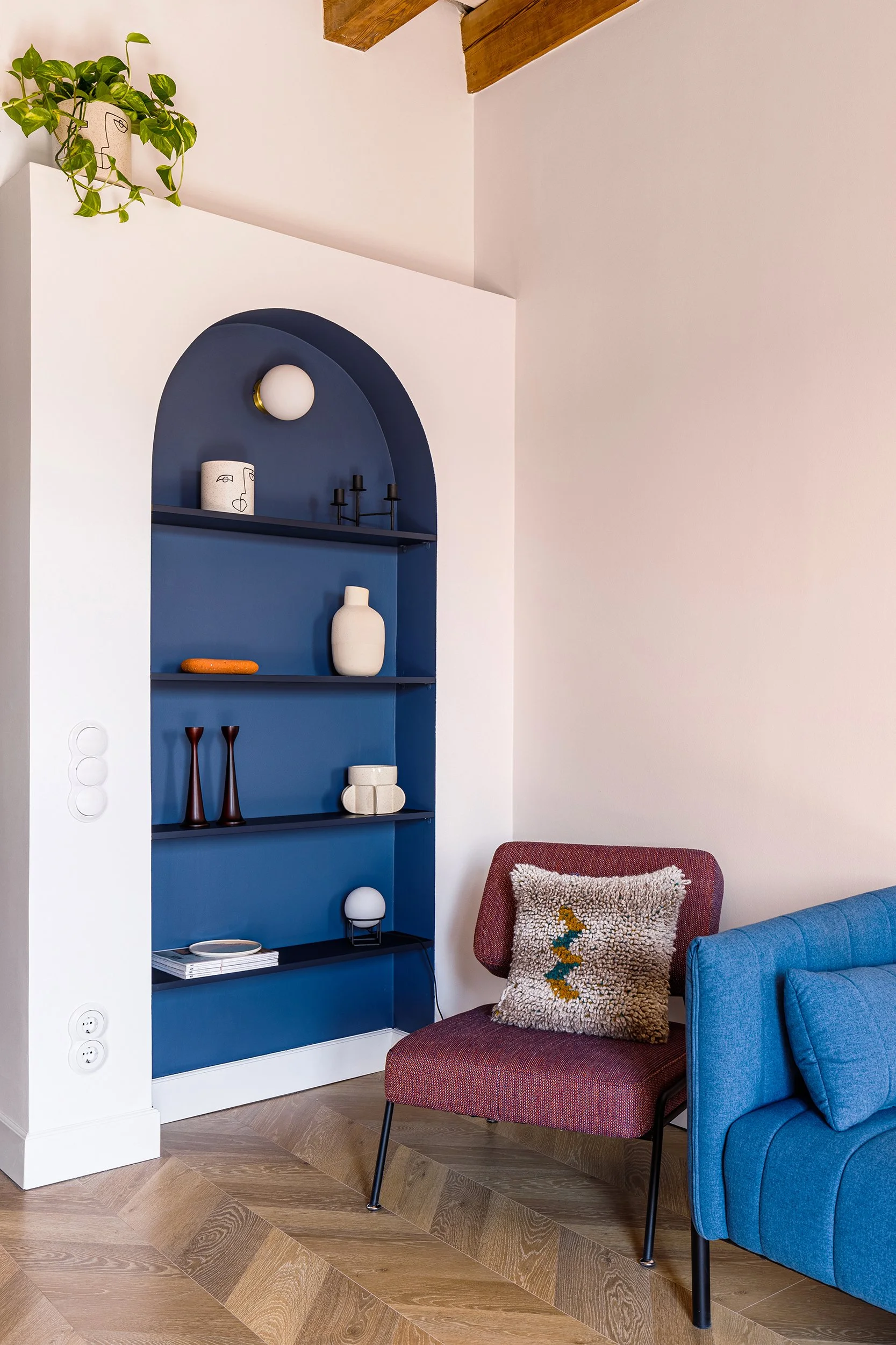 Interior with a blue built-in shelving alcove, featuring decorative vases, candles, and books. A plant is placed on top of the shelf. Below, a red chair with a textured cushion sits next to a blue sofa on a wood-patterned floor.