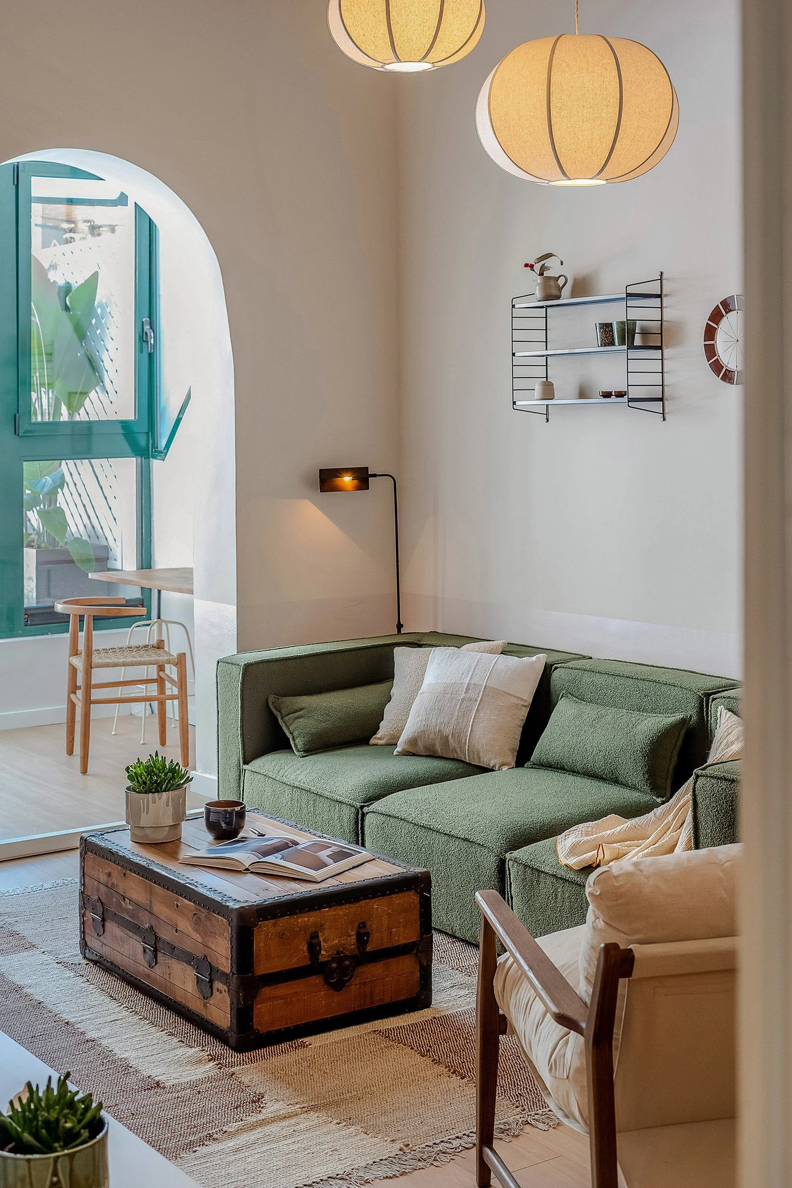 Interior of a cozy living room with a green sectional sofa, wooden trunk as a coffee table, and decorative plants. A wall-mounted shelf holds small items and a nearby window allows natural light to enter. Wooden chair and rug complete the setting.