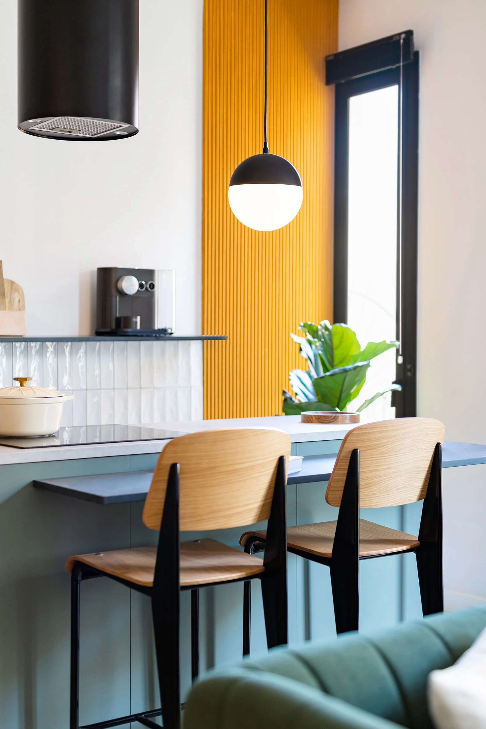 Modern kitchen interior with wooden chairs at a blue counter, pendant light, coffee machine, yellow accent wall, and green plant by the window.