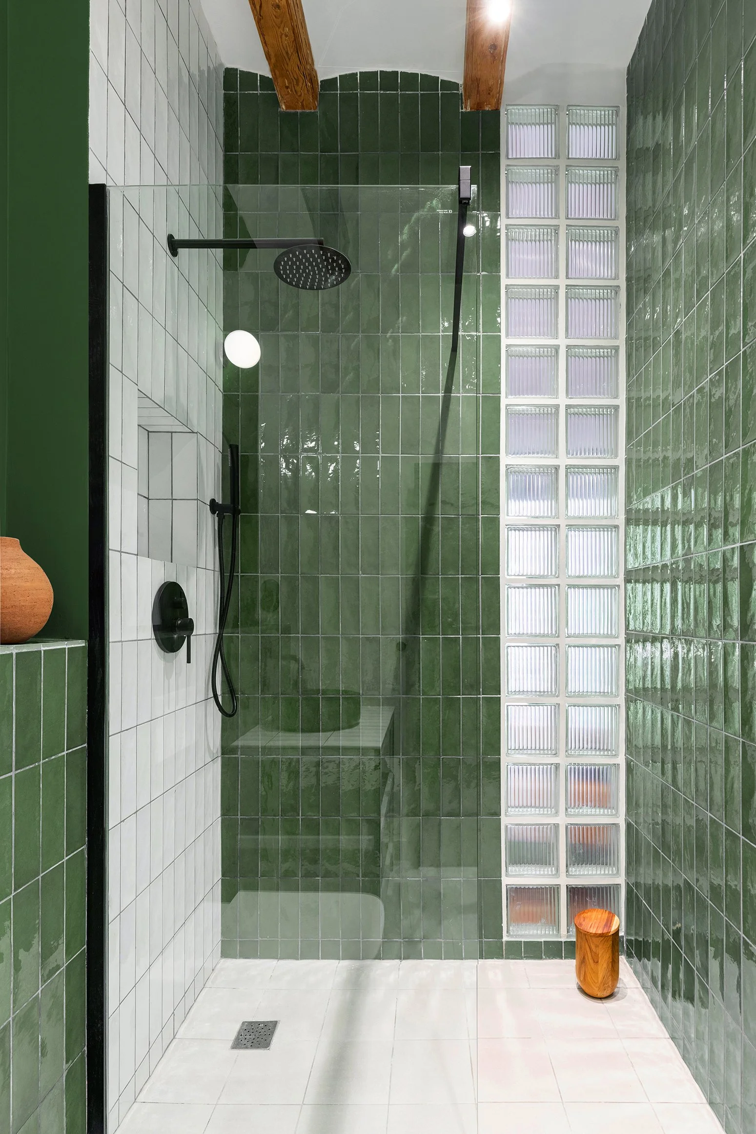 Modern shower with green and white tiles, glass wall divider, black showerhead, wooden stool, and exposed wooden beams.
