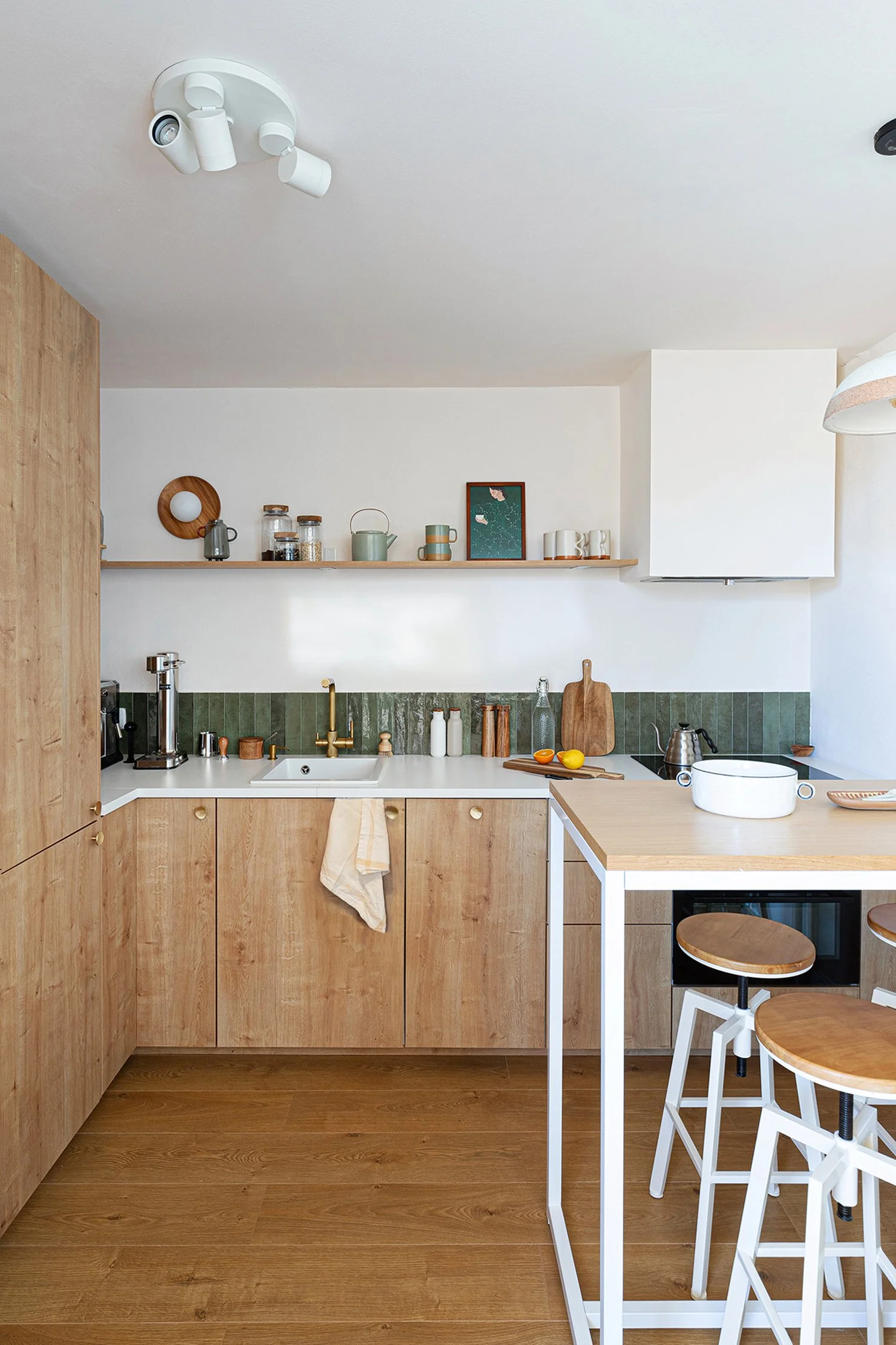 Modern kitchen with wooden cabinets, white countertop, bar stools, and open shelves. There are various kitchen items like a coffee maker, cutting board, and decorative jars. A towel hangs near the sink.