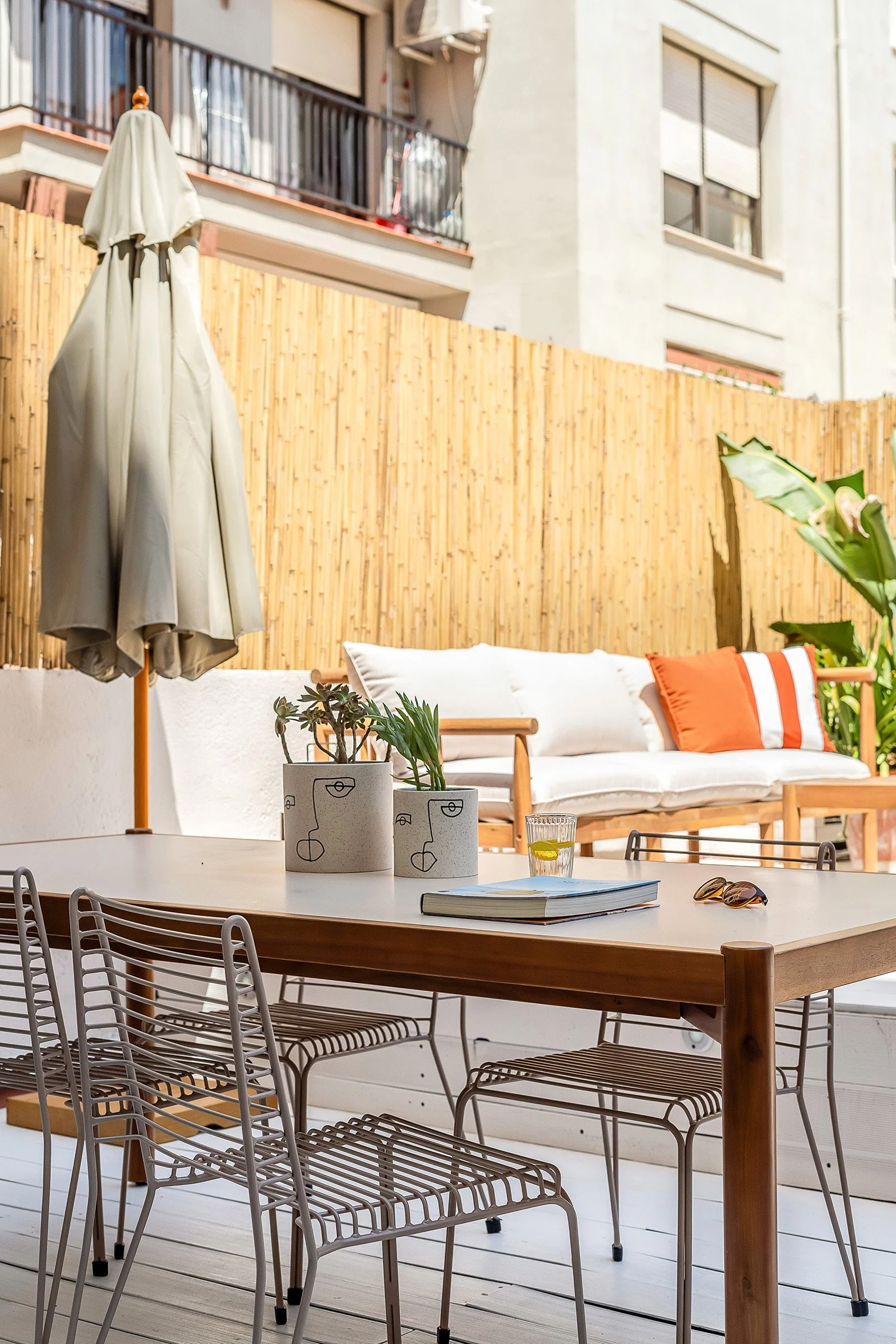 Outdoor patio with a wooden table, metal chairs, potted plants, closed umbrella, sofa with orange and white pillows, wooden fence, and nearby buildings.