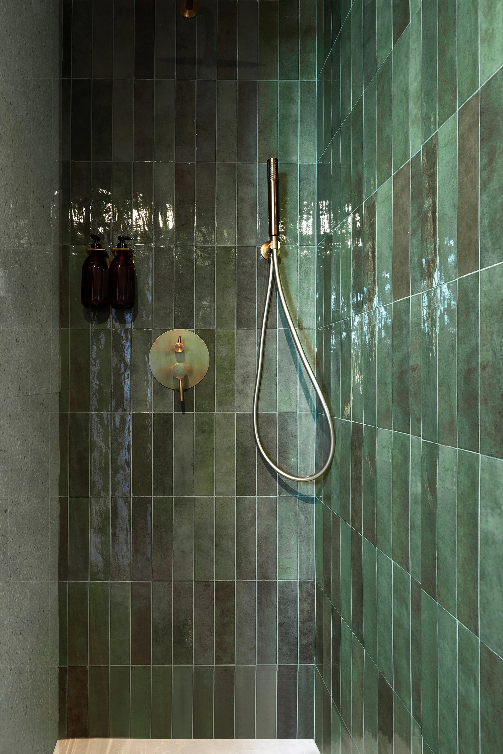 Modern shower with green tiles and brass fixtures, featuring a handheld showerhead and wall-mounted soap dispensers.