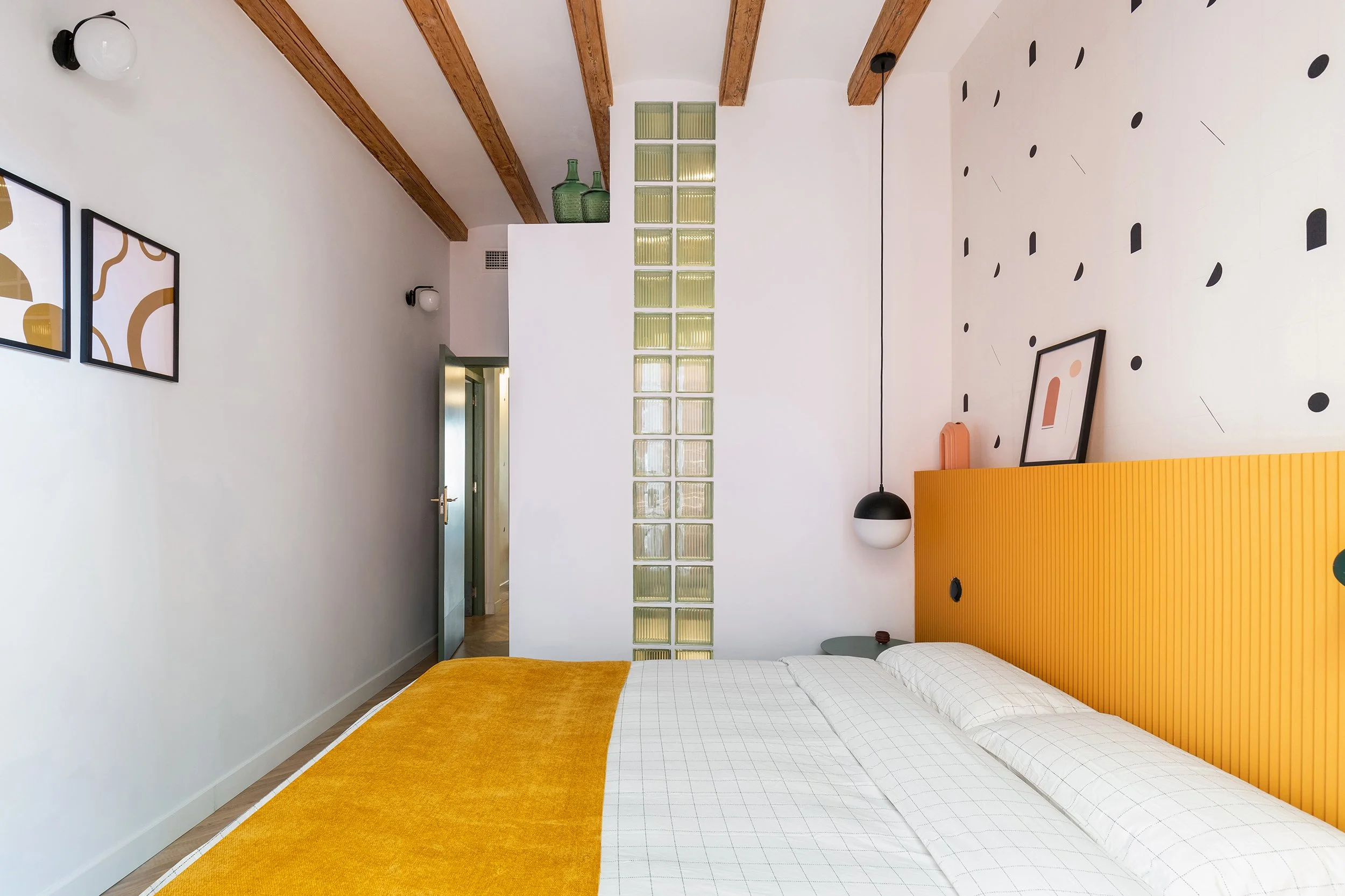 Modern bedroom with white walls, wooden beams on the ceiling, a yellow headboard, and a geometric-patterned wall. The bed has a yellow throw and checkered bedding. A glass block partition and minimalist decor, including wall art and pendant lighting,