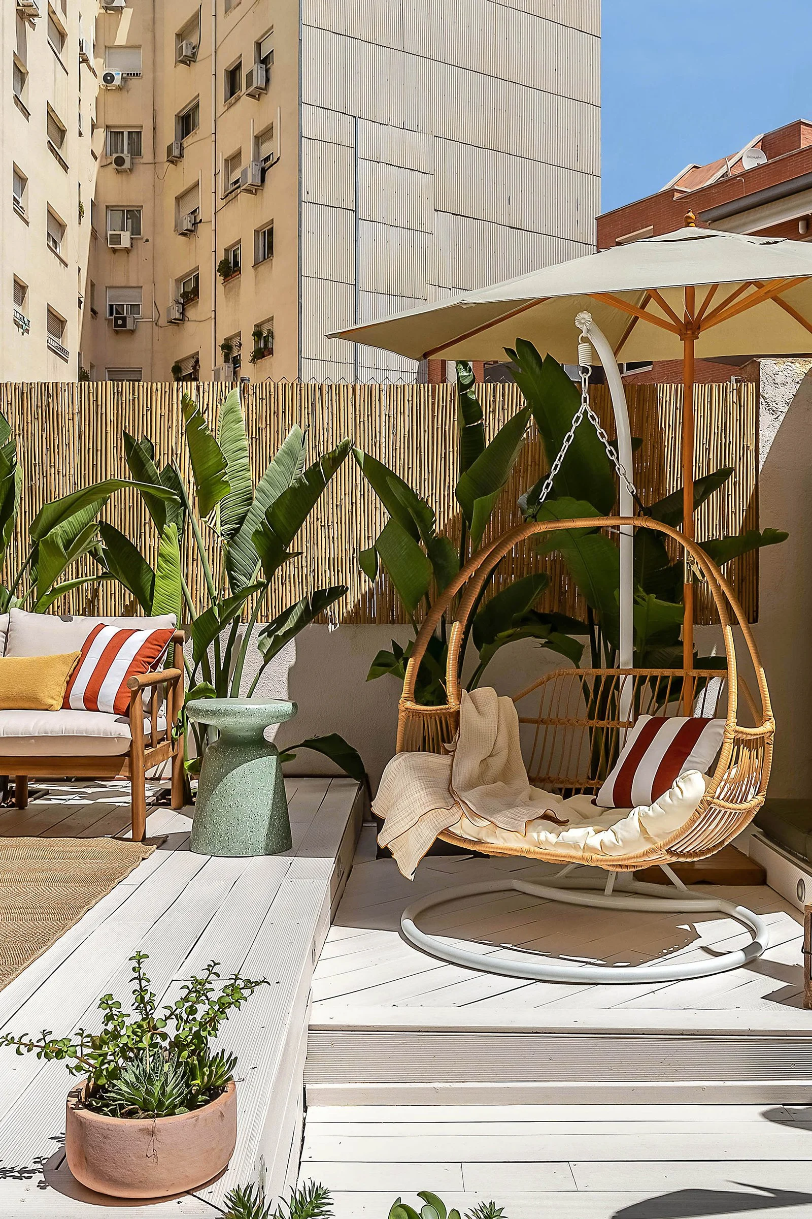 Outdoor patio with a rattan swing chair, striped cushions, a green side table, tropical plants, and a bamboo fence. An apartment building is in the background.