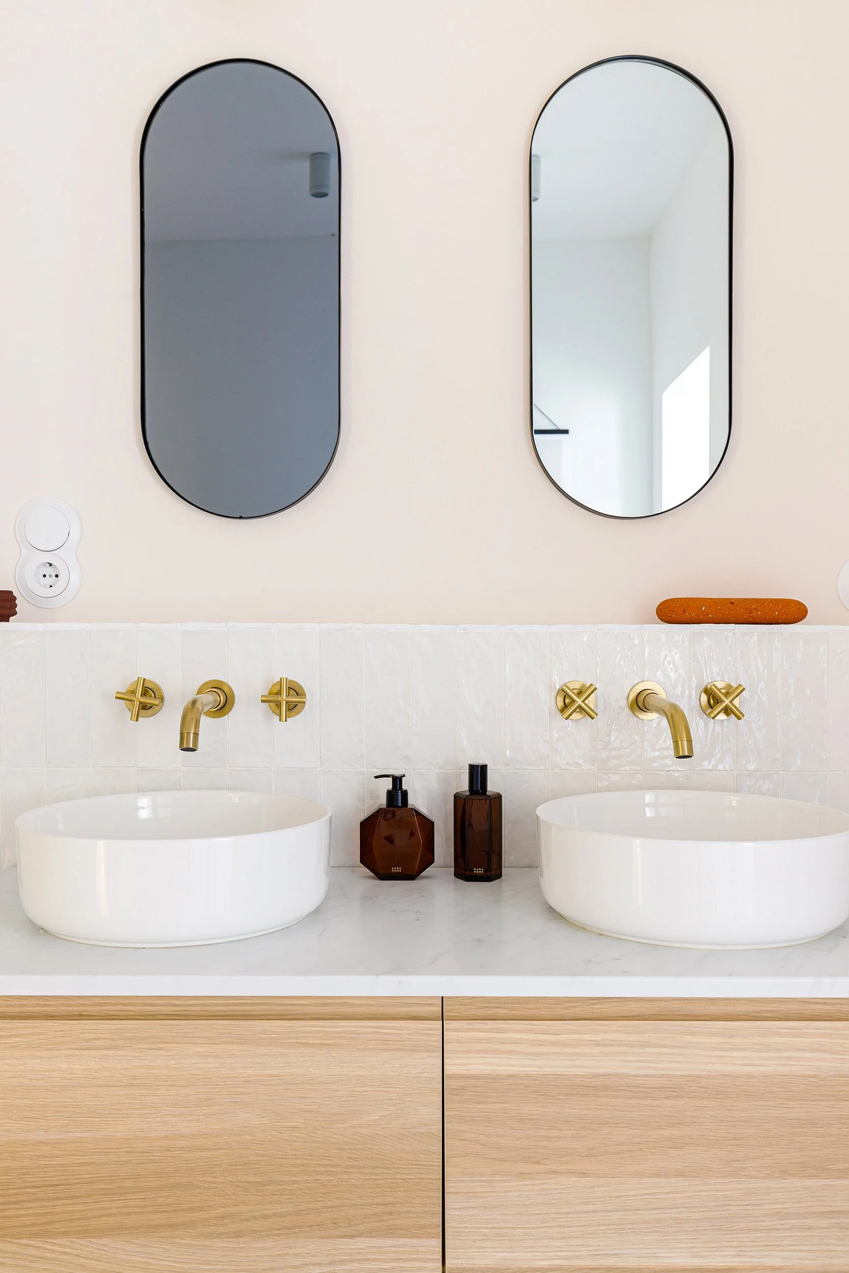 Modern bathroom with two white vessel sinks, gold faucets, and oval mirrors above each sink. Brown soap dispensers and containers are on the countertop, which is supported by light wood cabinetry.