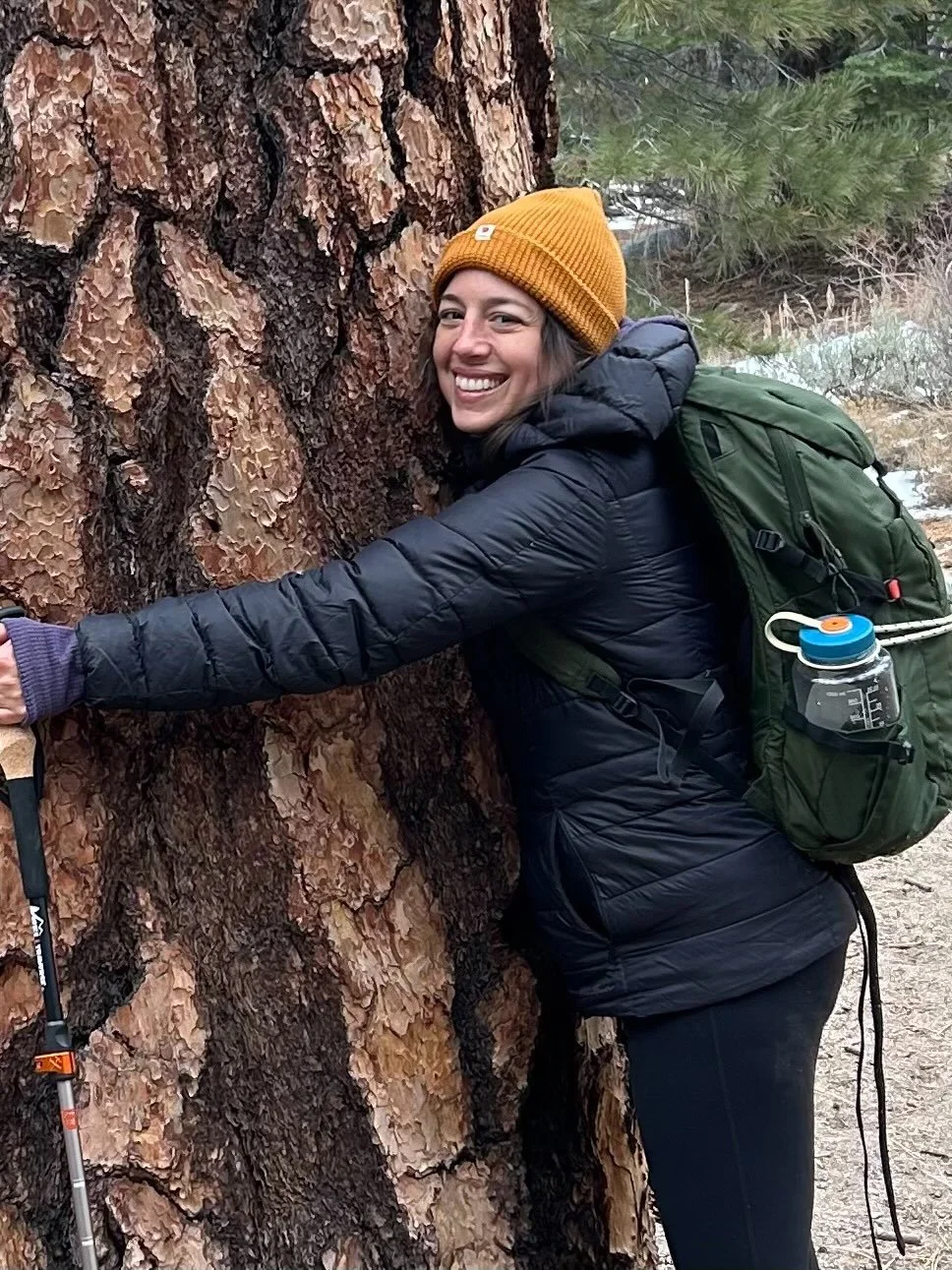 A woman hugging a large tree outdoors on a hiking trail, smiling and wearing a yellow beanie, black jacket, carrying a green backpack with a water bottle attached.