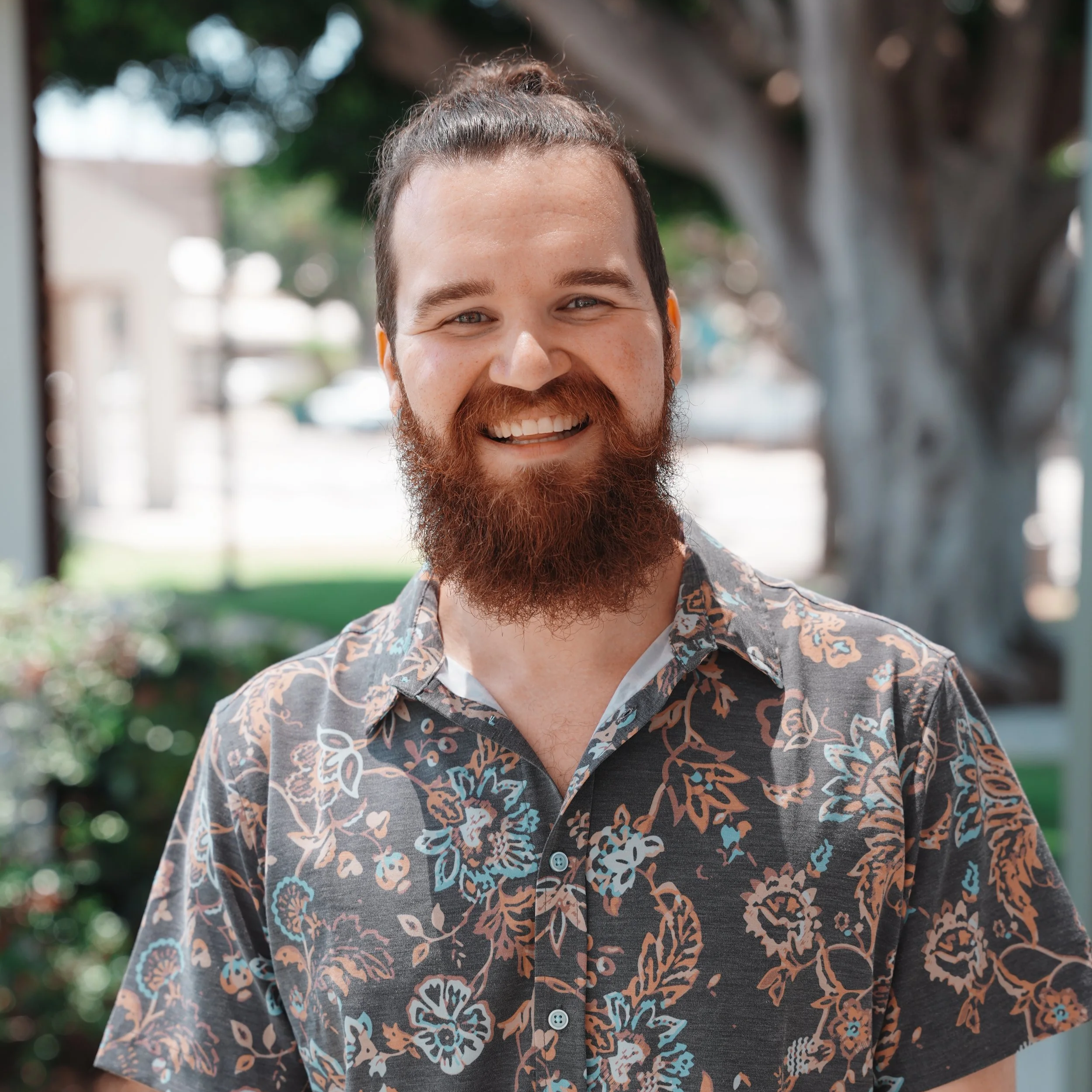 Close-up of a smiling man with a beard and mustache wearing a patterned shirt with floral designs, outdoors with blurry trees and sky in the background.