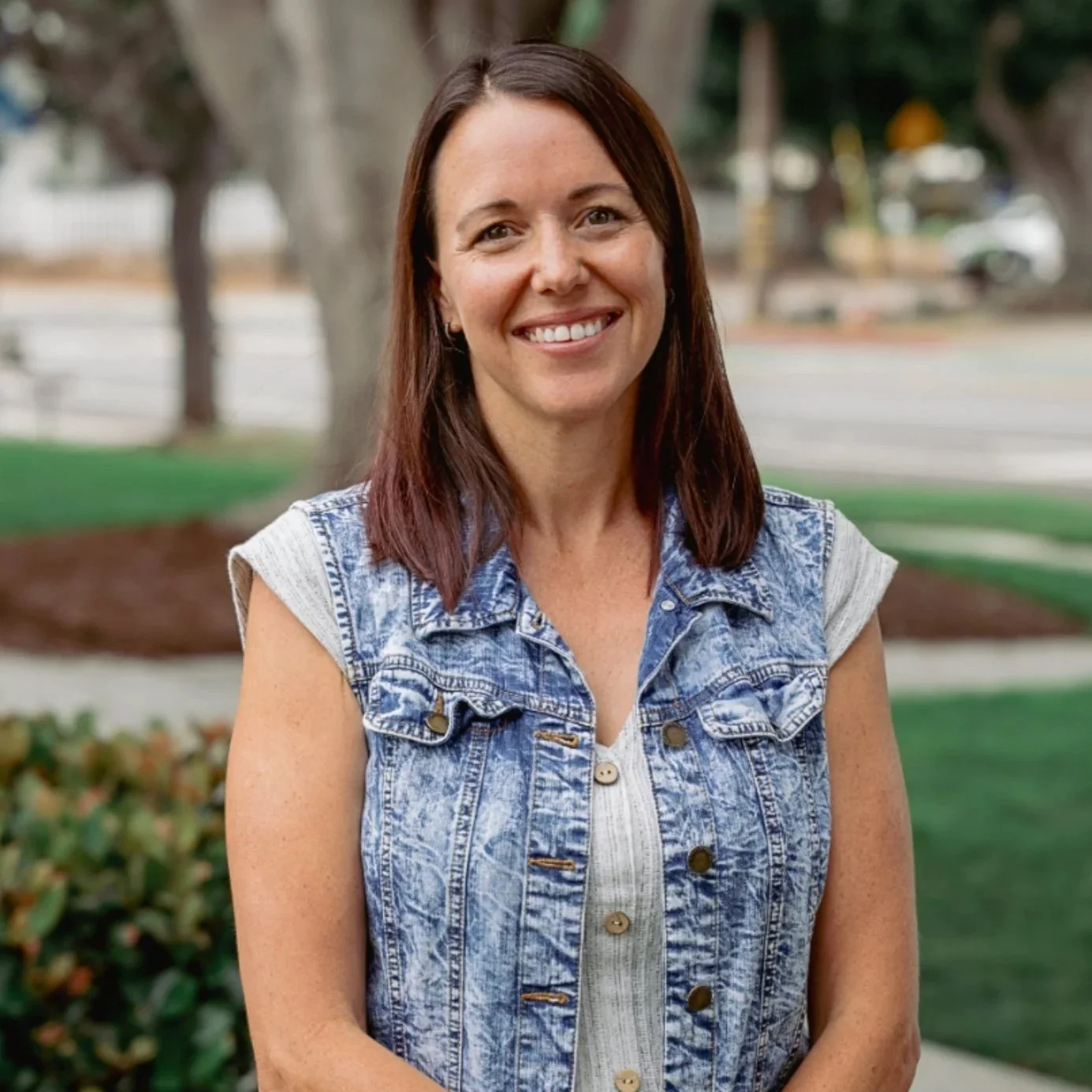 Portrait of a woman with brown hair and a smile, standing outdoors in a park with trees and greenery in the background, wearing a sleeveless denim vest over a white shirt.