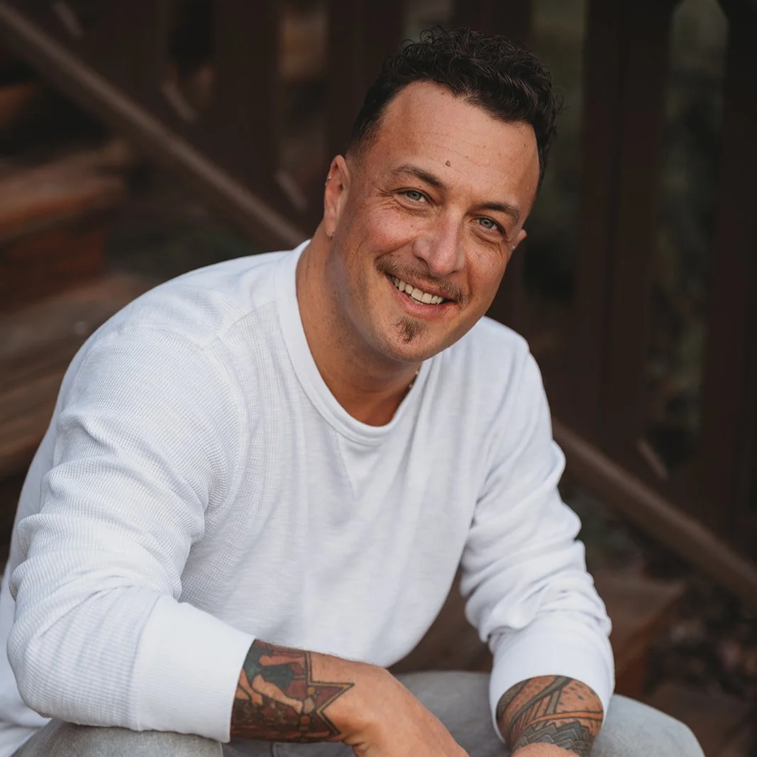 A smiling man with short dark hair, tattoos on his arm, wearing a white long-sleeve shirt, sitting outdoors near a wooden structure.