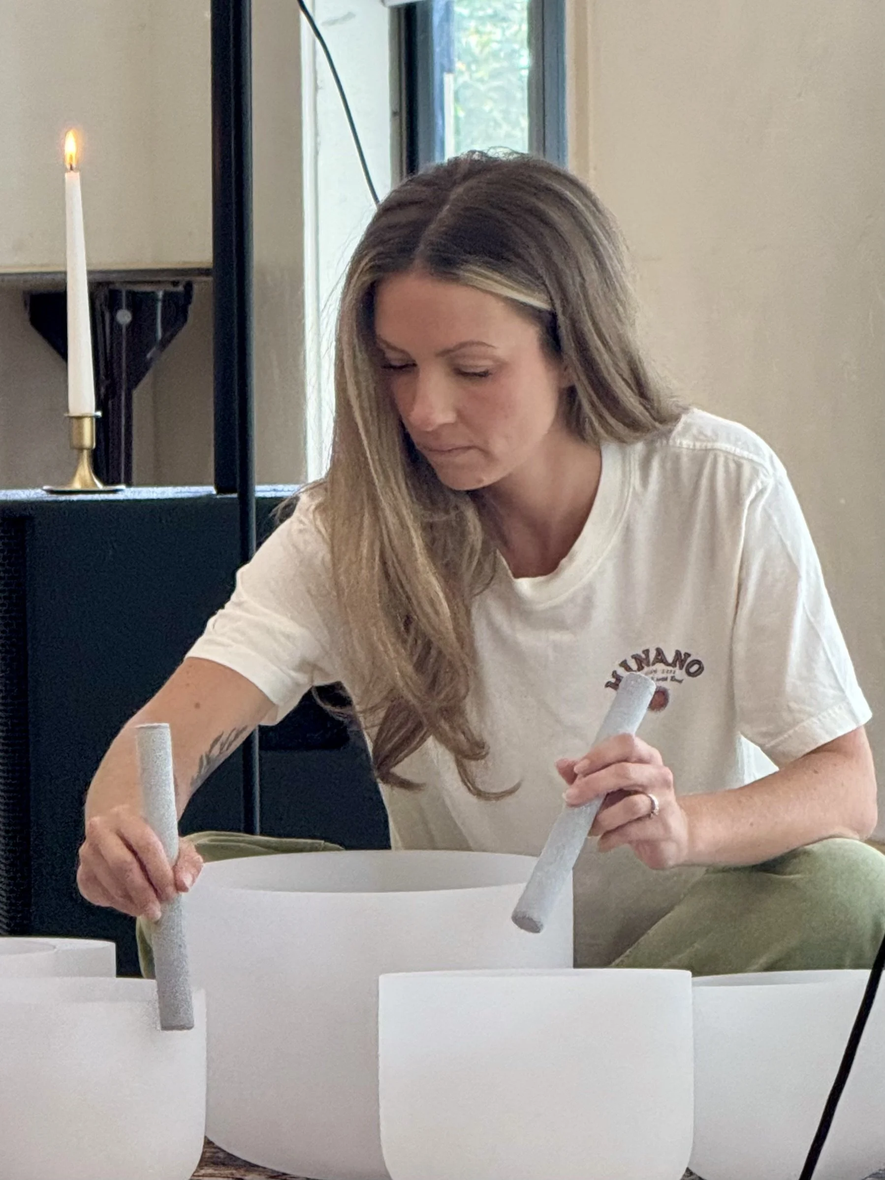 A woman with long, wavy, blonde hair and a white shirt with a logo is working on a white object, possibly a boat, with white tubes or pipes. She is in an indoor setting with a window in the background and a lit candle on a stand to her left.