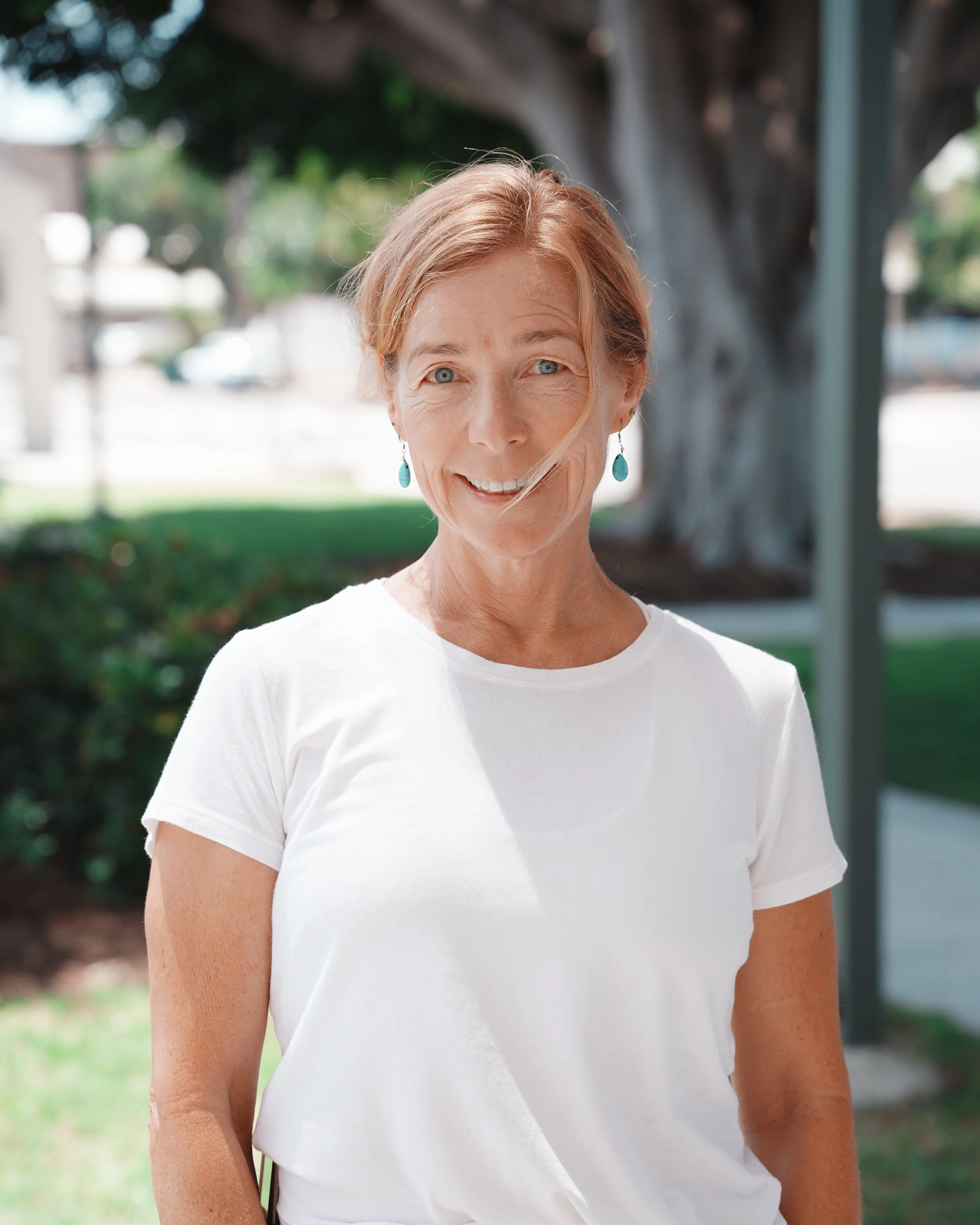 Close-up of a woman with short red hair, light blue eyes, wearing turquoise earrings, smiling outdoors with a blurry background of trees and a house.