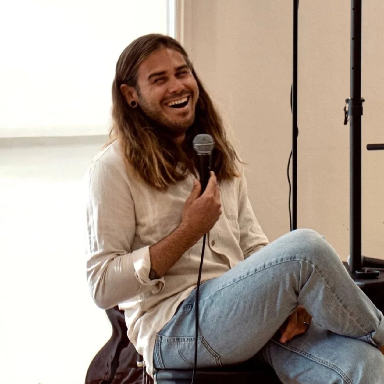 A man with long hair, wearing a cream-colored shirt and ripped jeans, sitting on a stool, smiling and holding a microphone during a performance. There is a large speaker, a microphone stand, and a hanging light in the background. The setting appears to be an indoor event space.