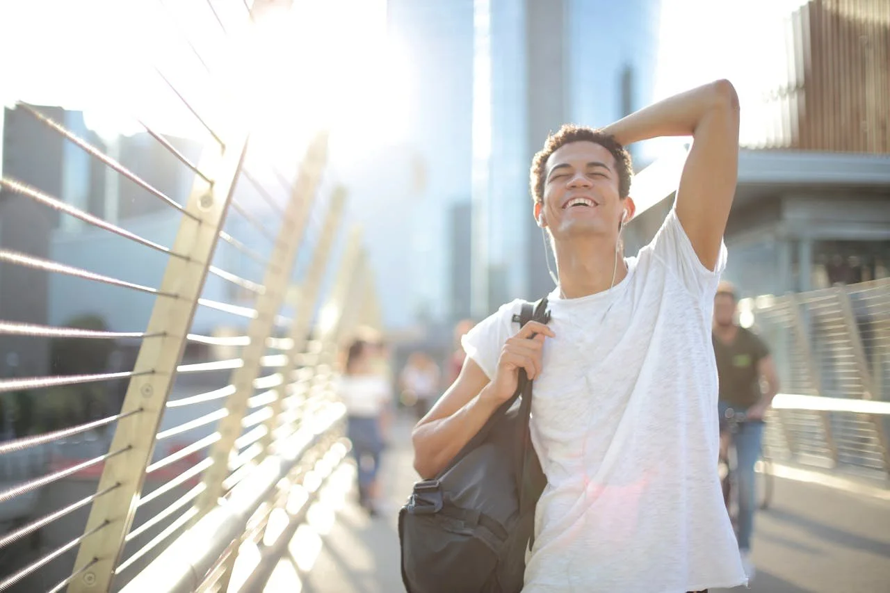 cheerful-ethnic-man-in-earphones-with-bag-on-shoulder-in-city-in-hot-summer-day