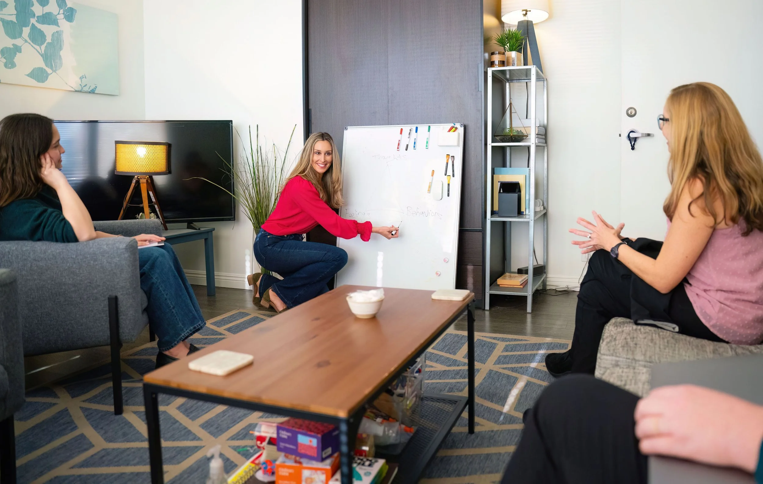 A woman in a red shirt is leading a presentation in a modern, cozy room with two other women seated and listening. There is a whiteboard with markers, a television, a lamp, and some shelves with plants and books, creating a professional yet relaxed atmosphere.