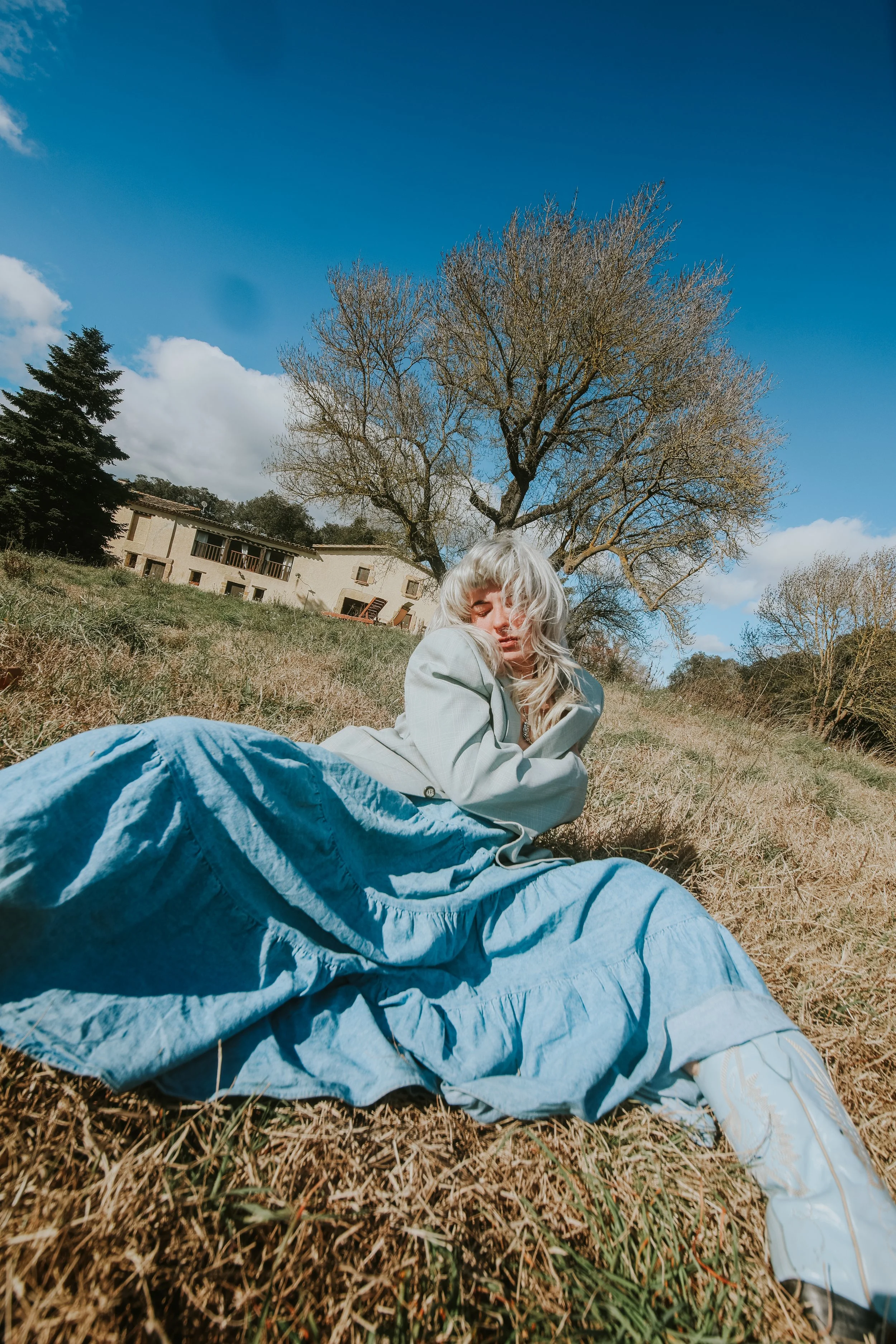a person with medium length, white blond hair sits with their legs wide. they are wearing a gray blazer, a long blue skirt, and blue cowboy boots. a large tree is in the background with an older spanish home further back. the sky is a deep blue.