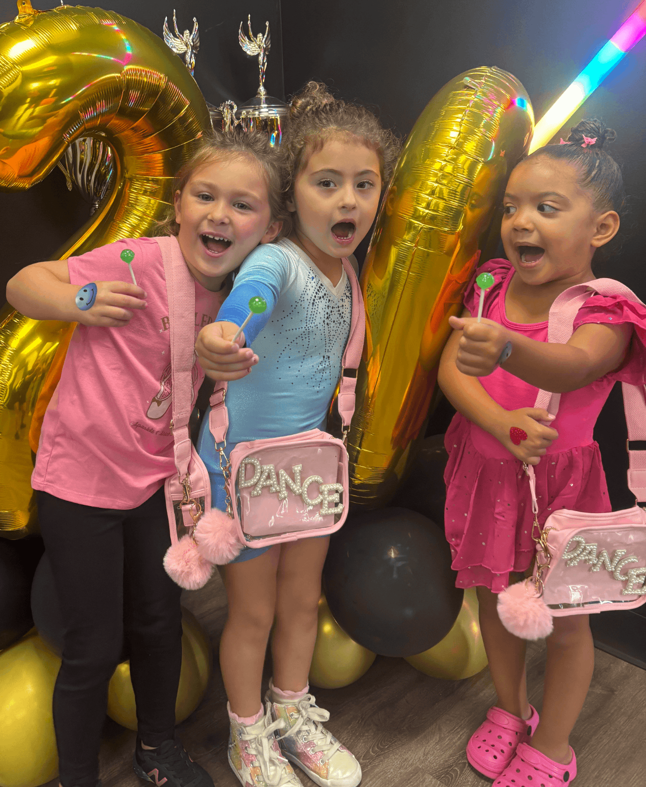 Three young girls celebrating a birthday with balloons, some shaped like the number 4, and balloons with the word 'DANCE' on their bags, in front of a black wall with decorative silver ornaments.