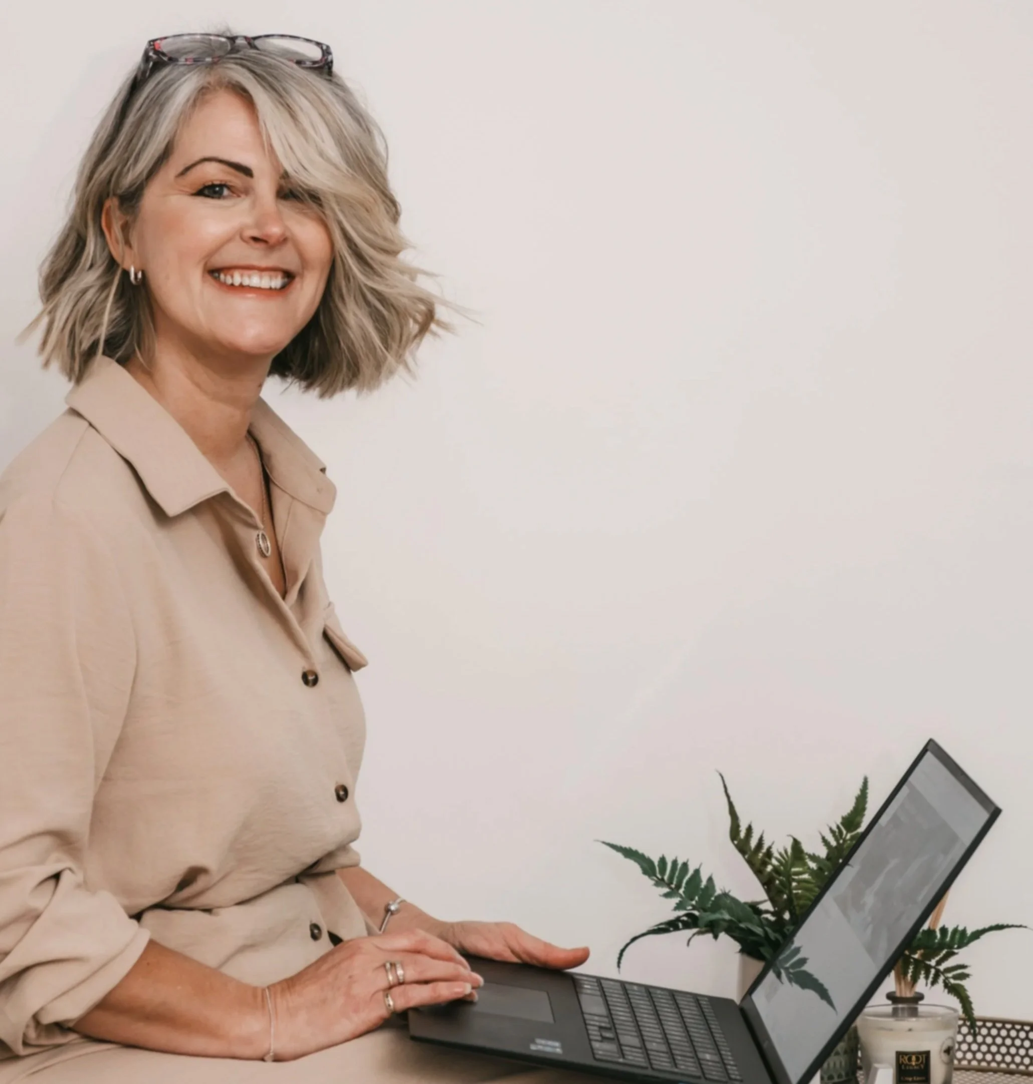 A smiling woman with blonde, shoulder-length hair, wearing glasses on her head, working on a laptop at a desk with a potted plant, candle, and a white background.