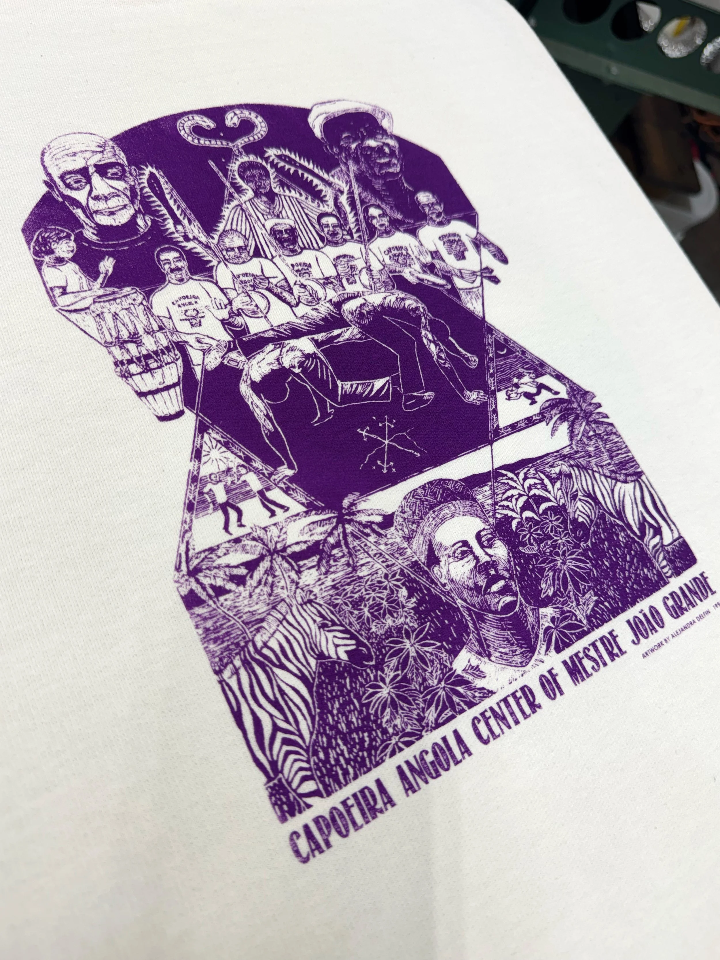 Purple illustration of a group of indigenous people, with a large face of a man and other figures, surrounding a decorated structure, with text at the bottom reading 'CAPOEIRA ANGOLA CENTER OF MESTRE JOAO GRANDE'.