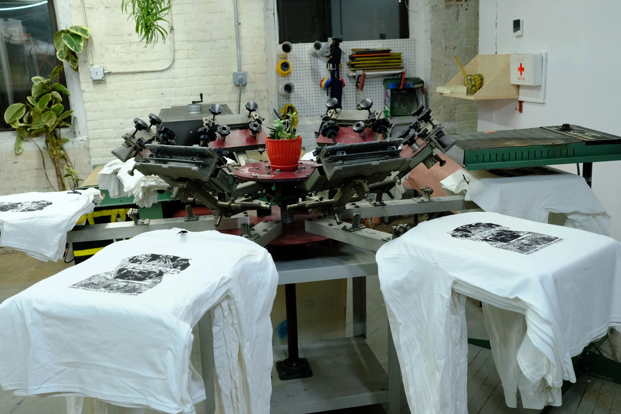 Silkscreen printing equipment with white T-shirts featuring black printed designs laid flat on tables in an art studio or workshop.