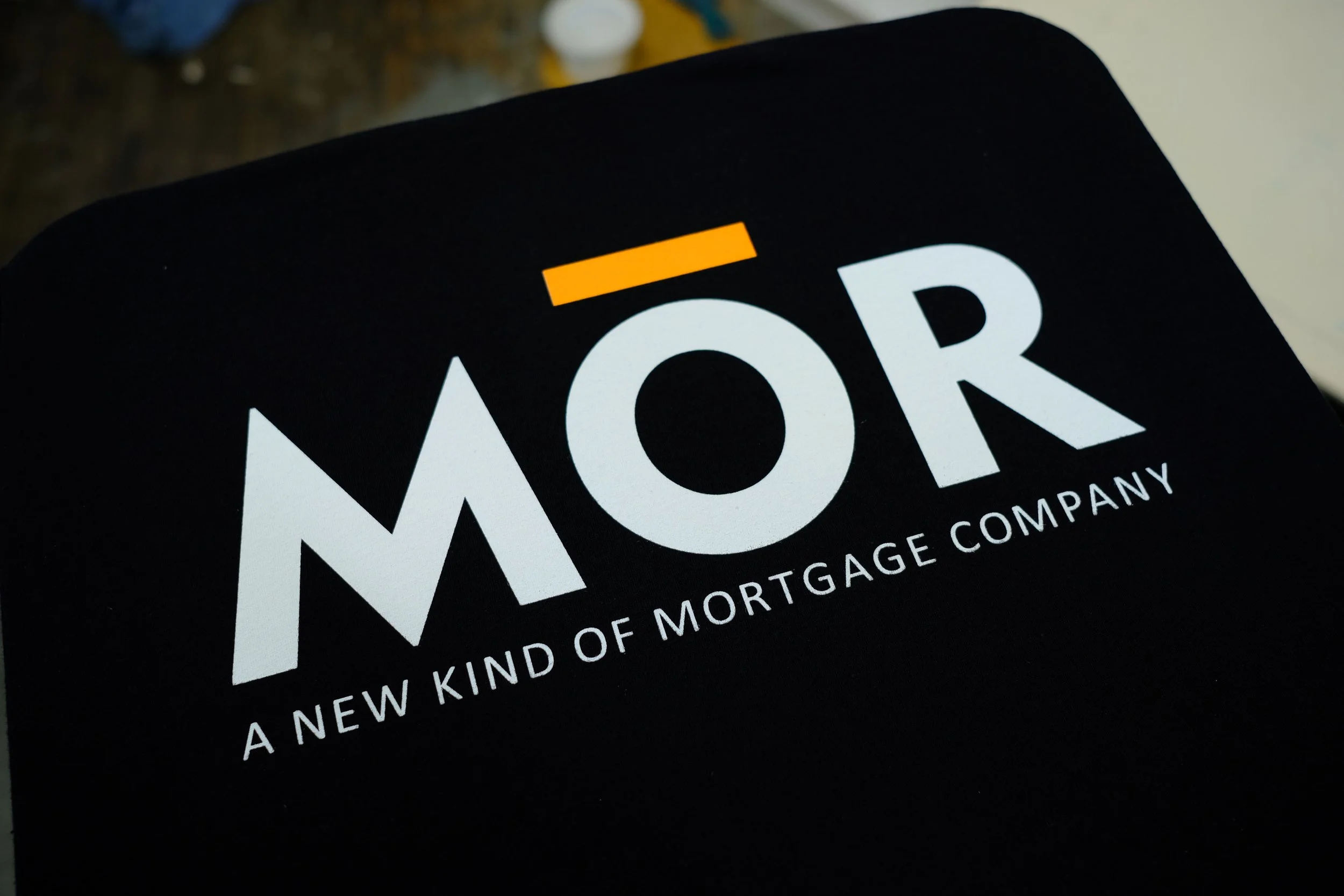 Close-up of a black sign with white text reading 'A NEW KIND OF MORTGAGE COMPANY' and a yellow accent above the letter 'O' in 'MOR'. The background shows a blurry floor with some objects.