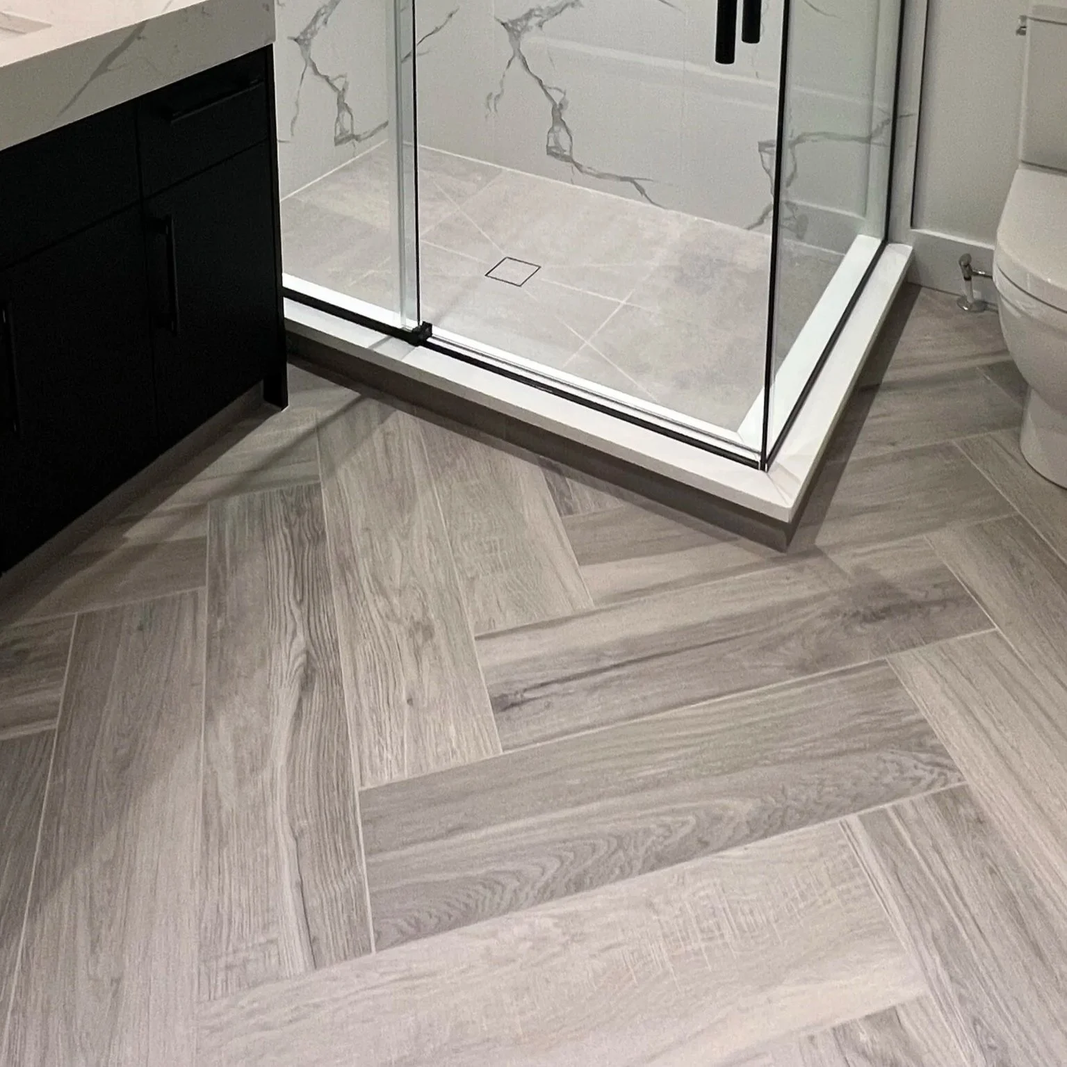 Bathroom with beige and gray wood-look tile flooring, a glass shower with marble walls, black cabinetry, and a white toilet.