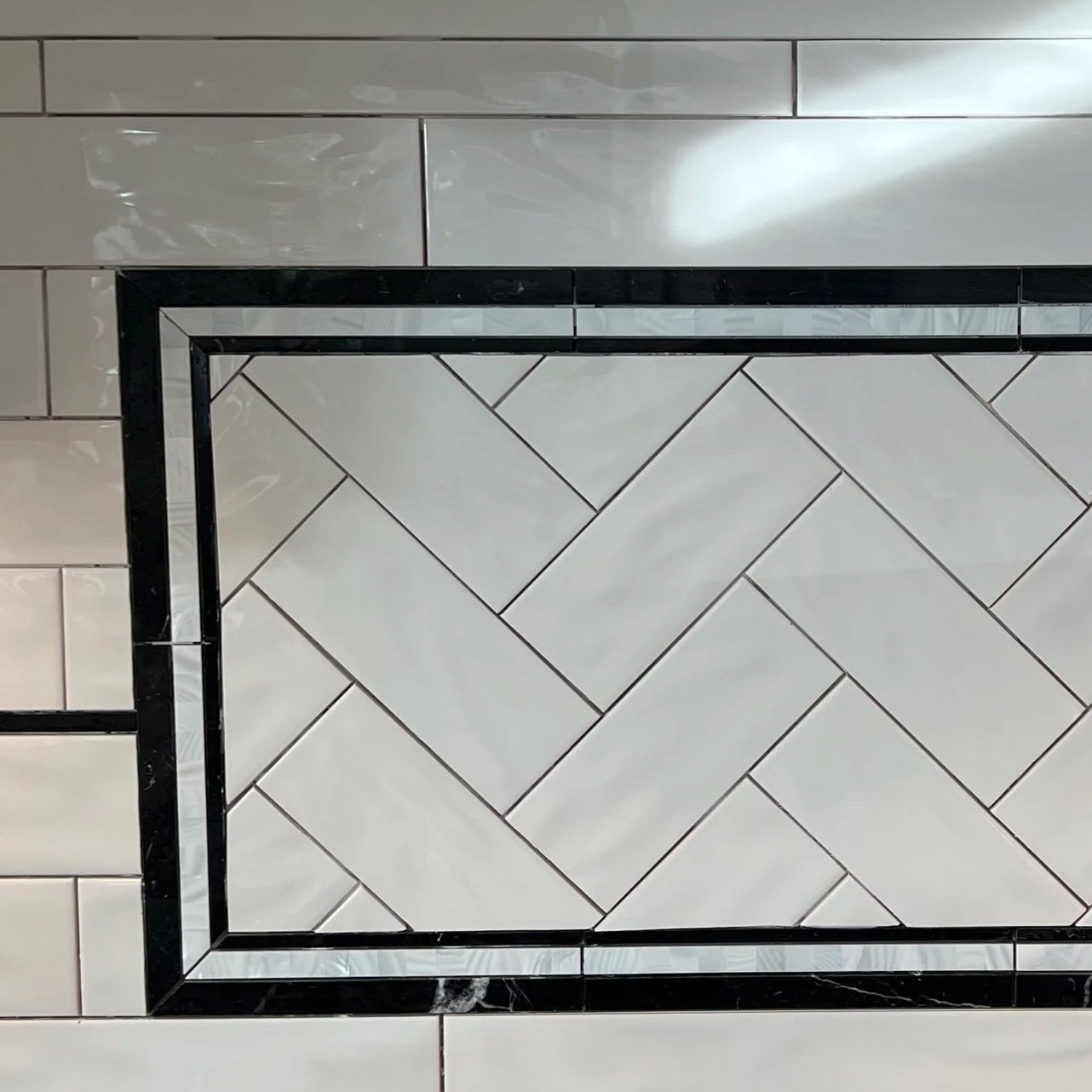 Top view of a tiled floor section with white and black tiles. The larger section has white tiles arranged in a herringbone pattern, bordered by a black tile frame.
