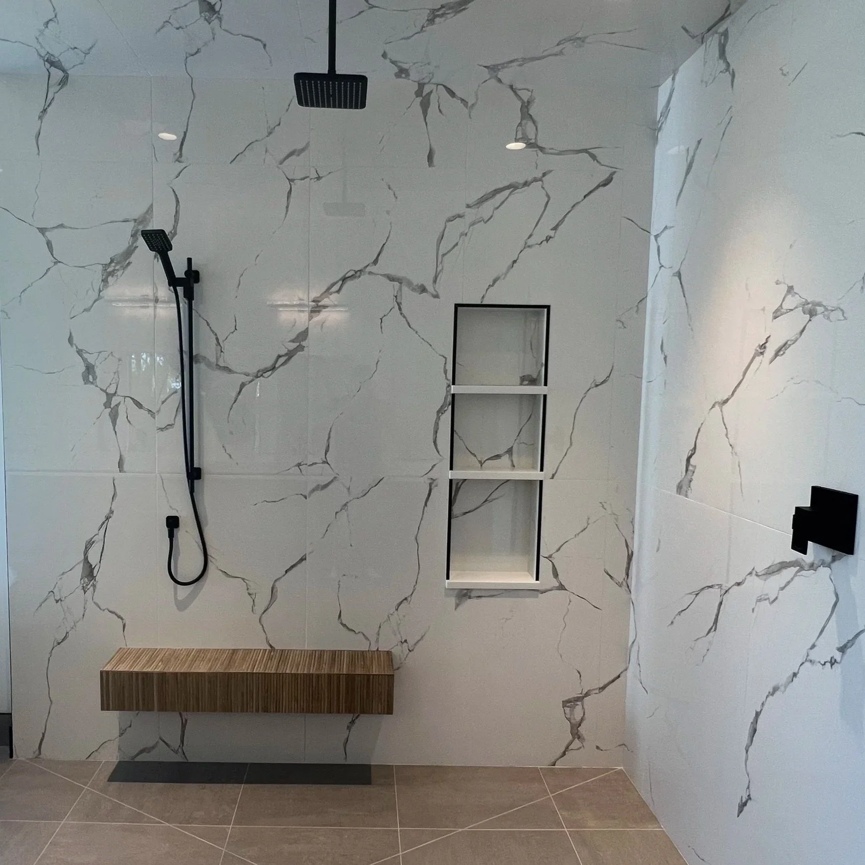 Modern shower with white marble walls with gray veining, black shower fixtures, a wooden bench, a white wall-mounted shelf, and beige tiled floor.