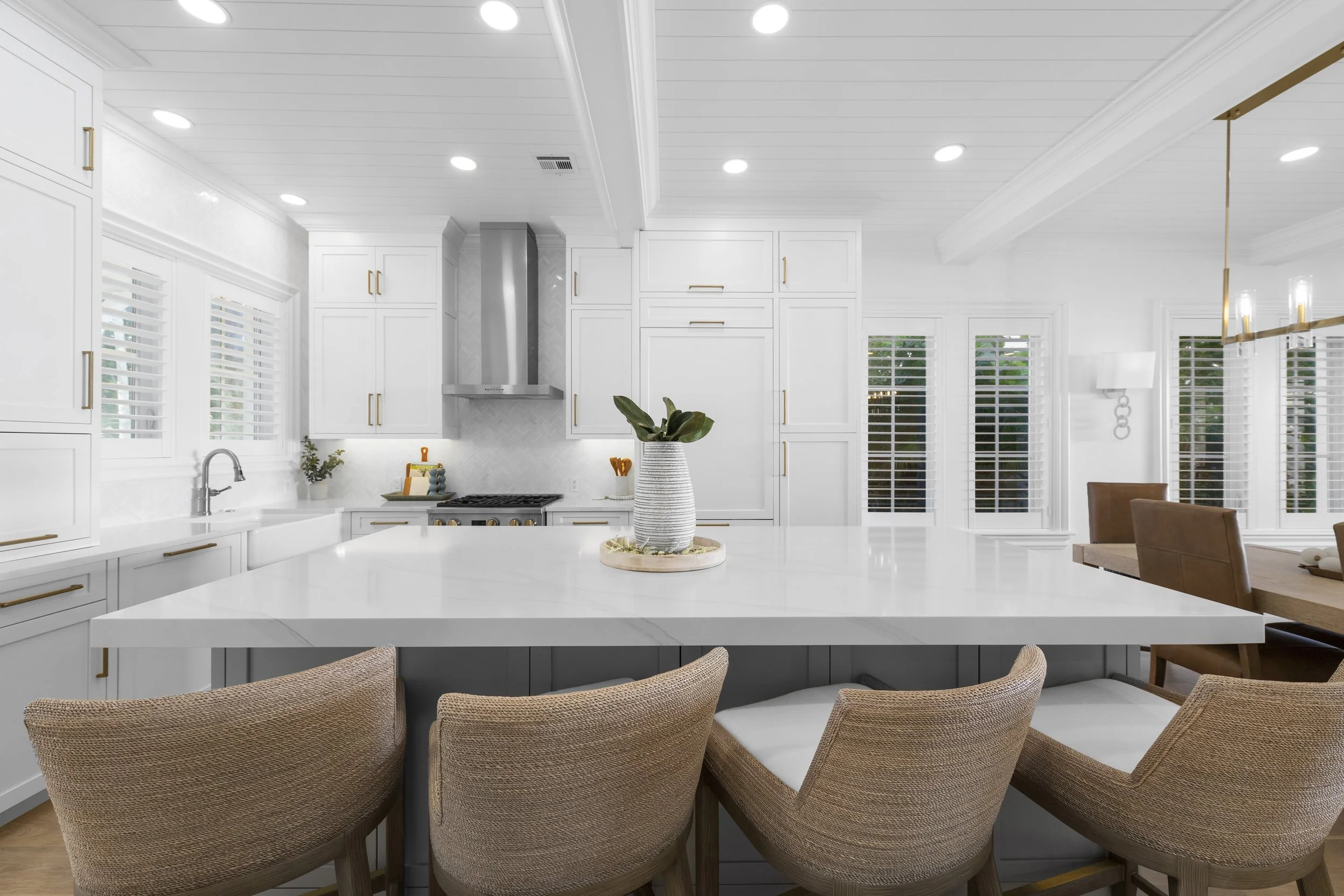 Bright white kitchen with a large central island, beige bar stools, and white shiplap ceiling. There are gold handles on white cabinets, a stainless steel hood above a stove, and multiple windows with white blinds.
