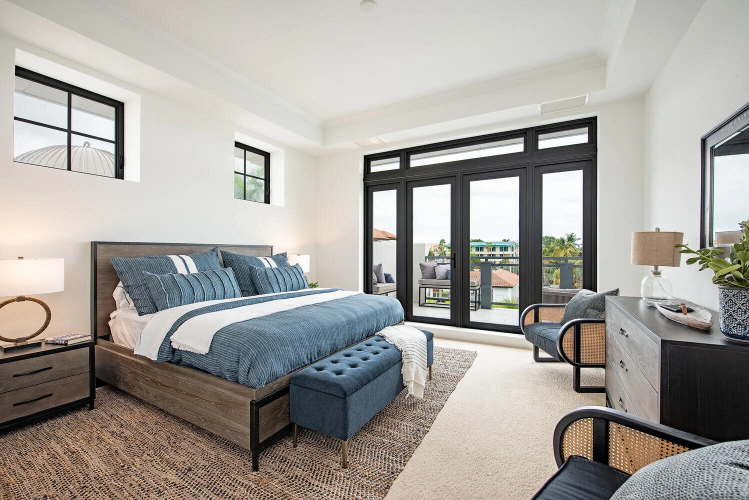 Modern bedroom with a large bed, blue bedding, bedside lamps, wicker side chair, black dresser with decorative items, and a balcony with outdoor seating and a view of buildings and palm trees.