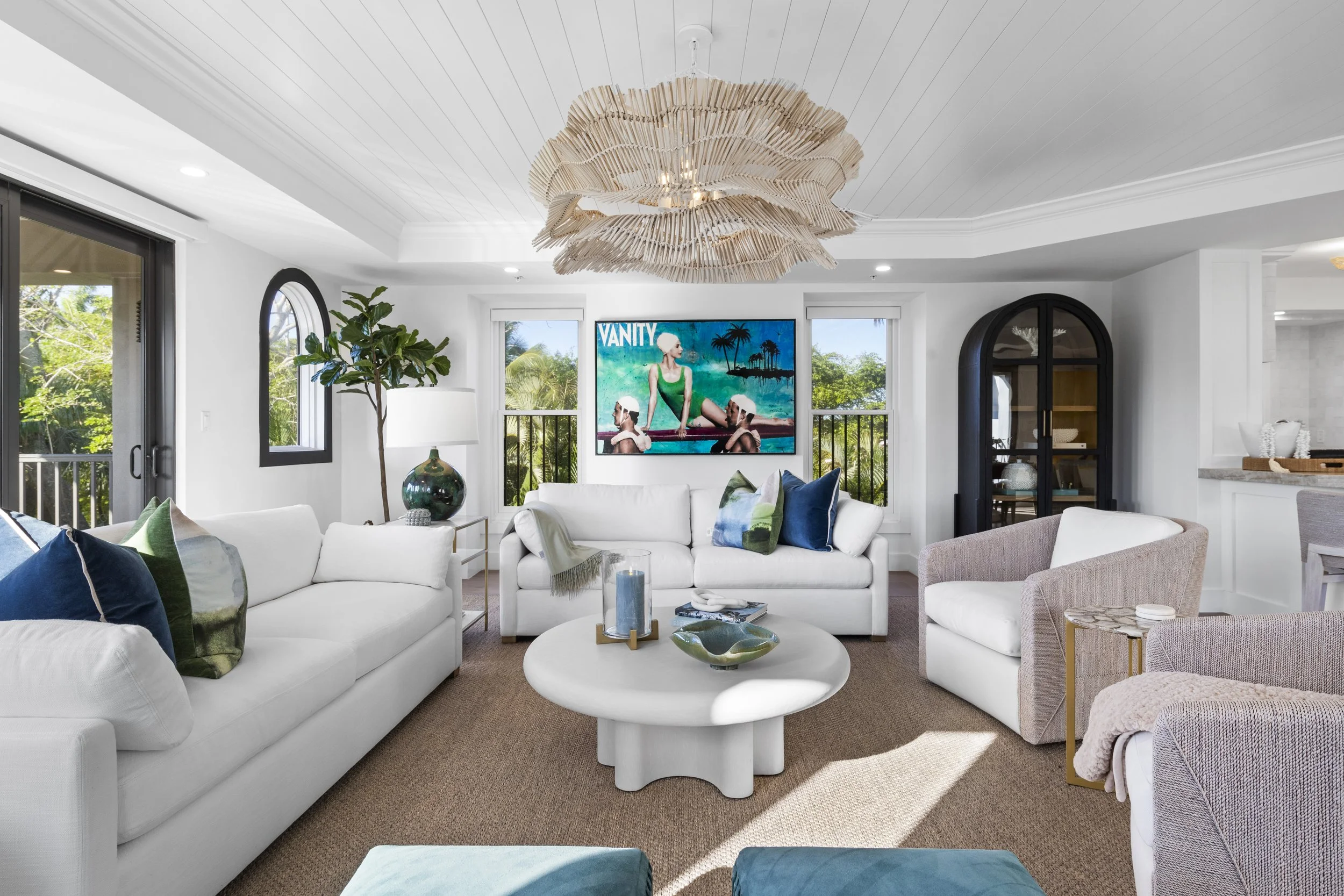 Living room with white sofas, colorful pillows, a large circular white coffee table, a tall potted plant, a modern chandelier, large windows with views of greenery, and a flat-screen TV on the wall.