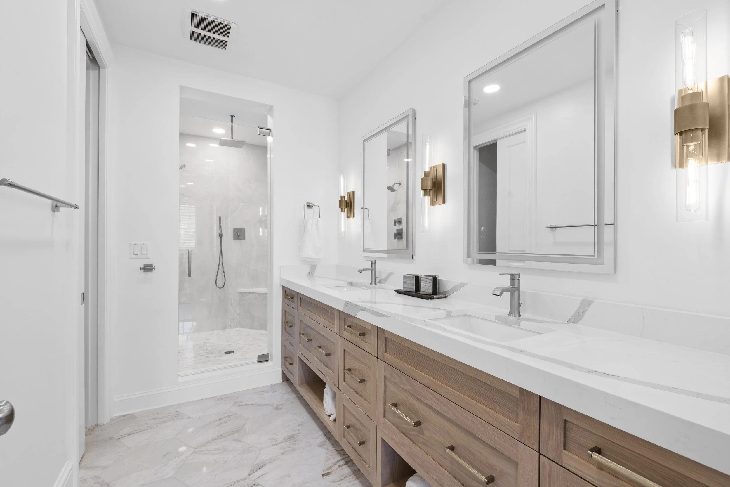 Modern bathroom with a double vanity featuring white marble countertops, two mirrors, gold fixtures, and a glass-enclosed shower with a rainfall showerhead and marble walls.