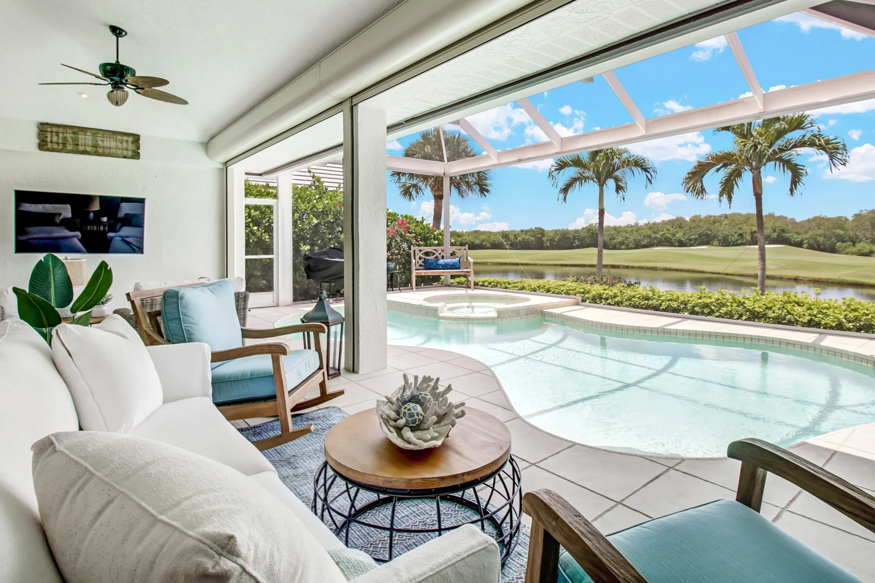 Living room with white sectional sofa, wooden chairs with blue cushions, and a coffee table with a decorative coral centerpiece, overlooking a screened pool area with palm trees, water view, and a bright blue sky.