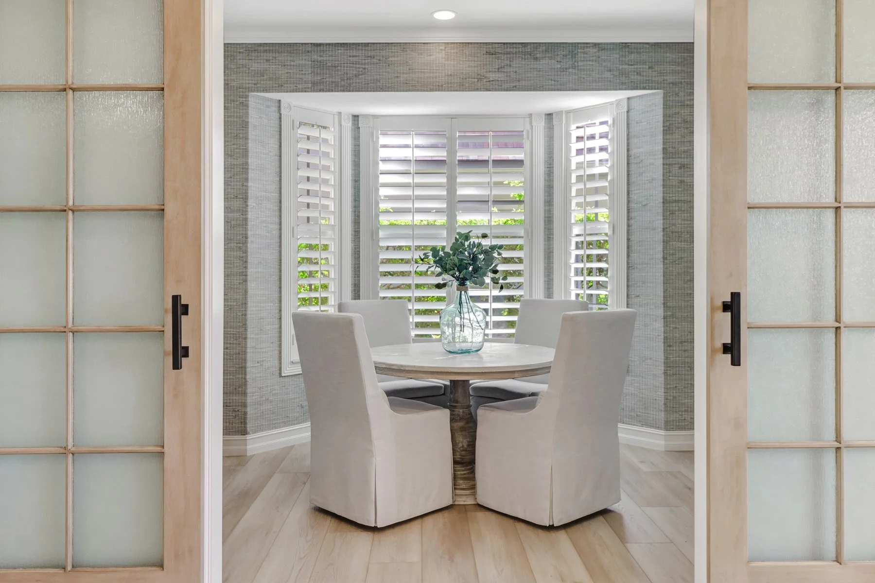Dining room with a round table and six white upholstered chairs, a vase with greenery, and a bay window with white plantation shutters in the background.