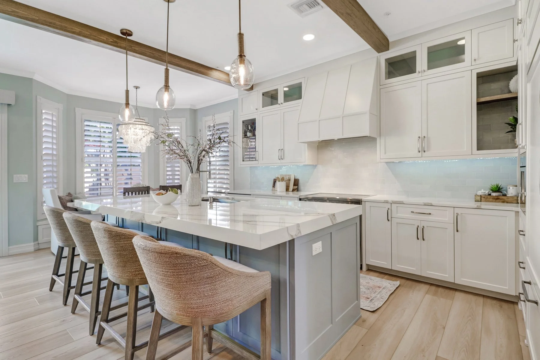 Modern kitchen with white cabinets, marble island, wood beams, and light wood flooring, with seating area and large windows with shutters.