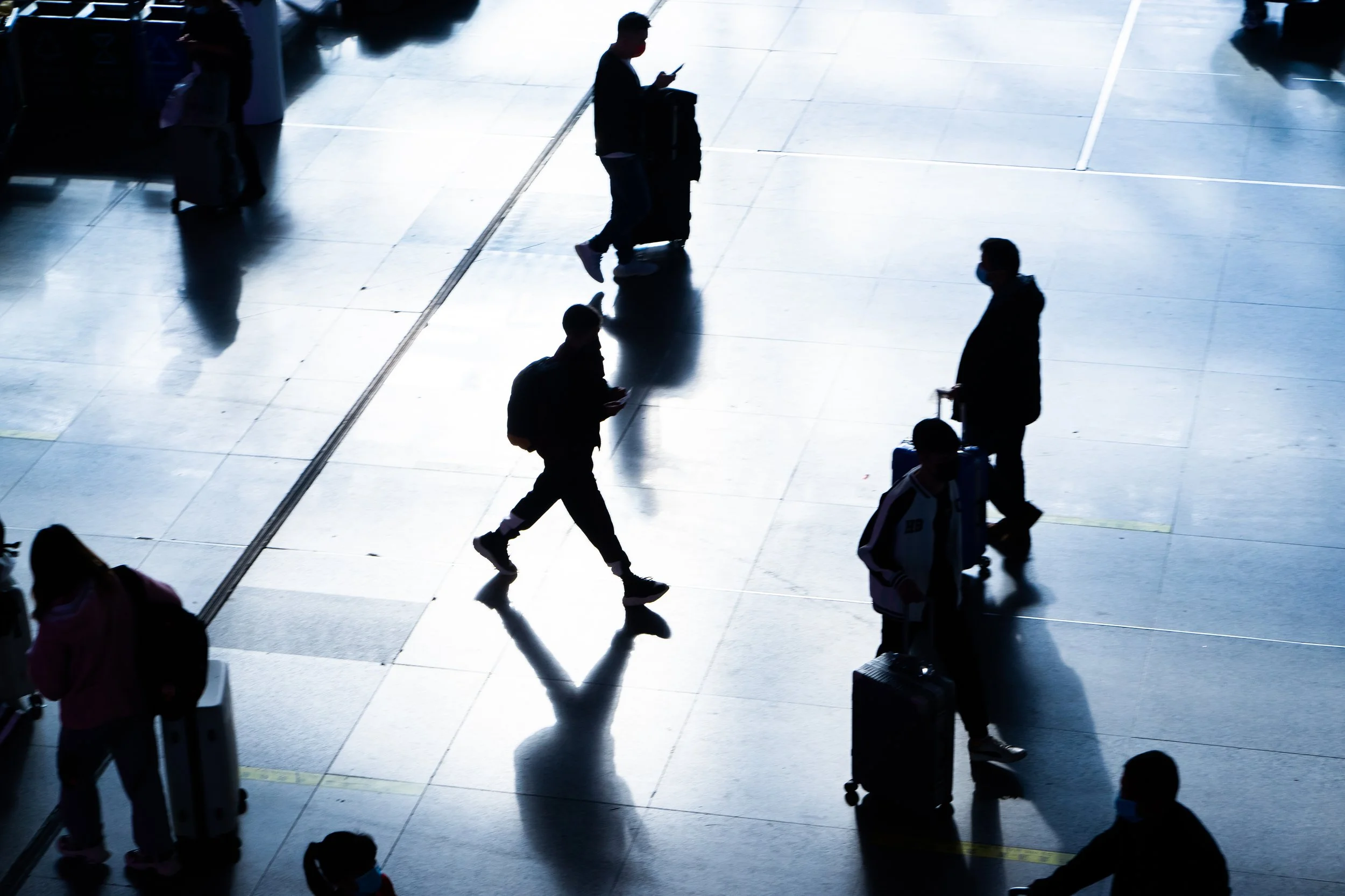 Silhouettes of travelers walking through an airport terminal with their luggage and some looking at their phones.