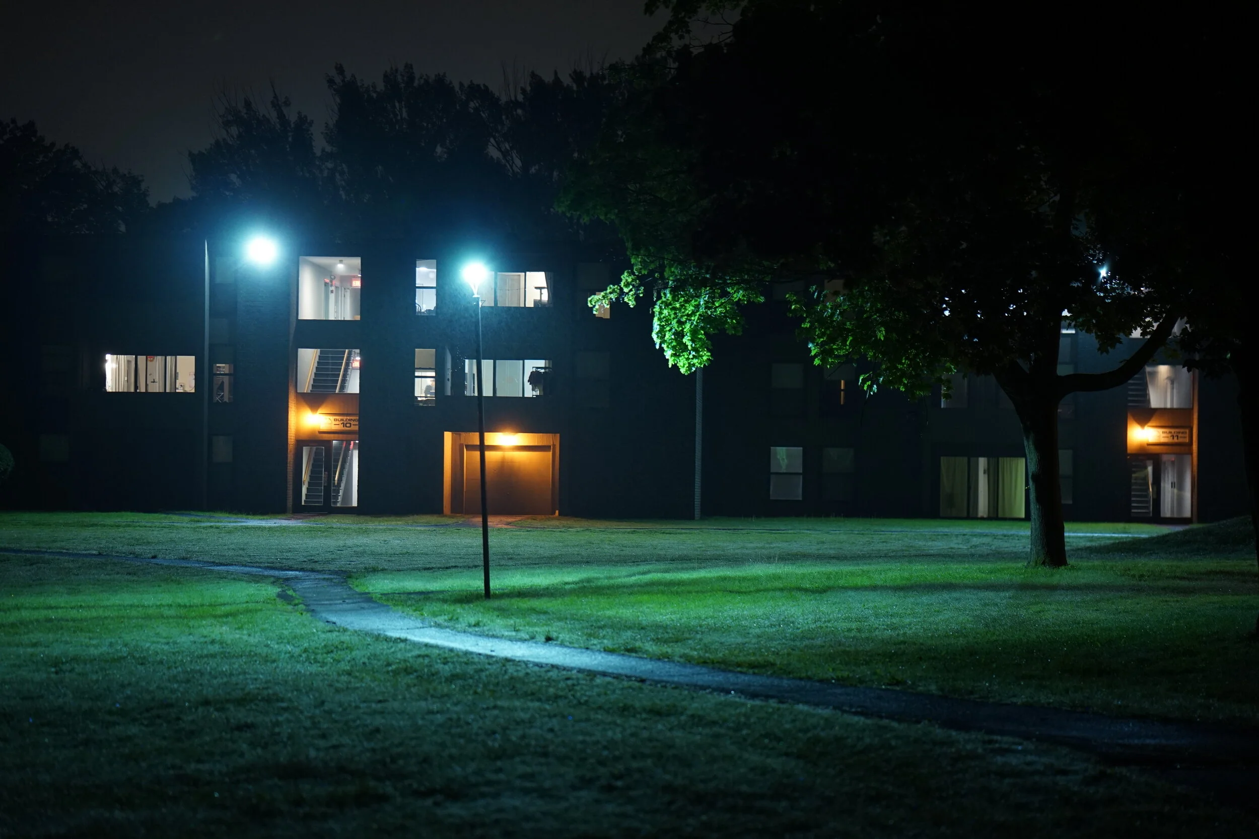 Nighttime view of a modern apartment building with lights on inside, surrounded by a grassy area and trees.