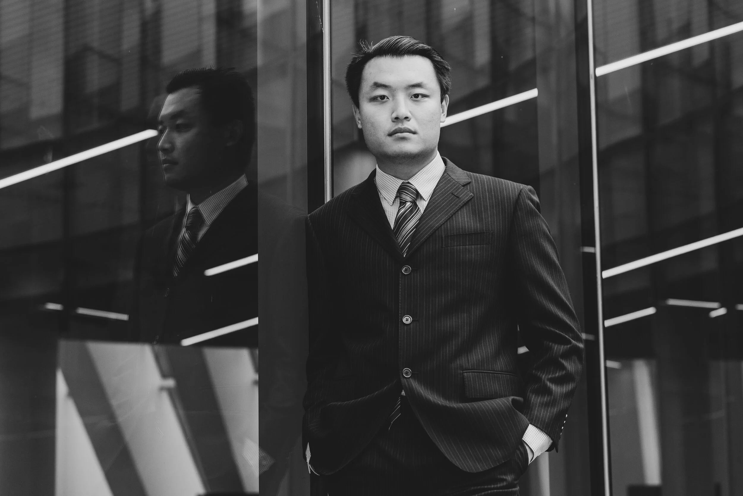 Black and white photo of a man in a business suit standing with his hands in his pockets next to a glass wall, with his reflection visible.