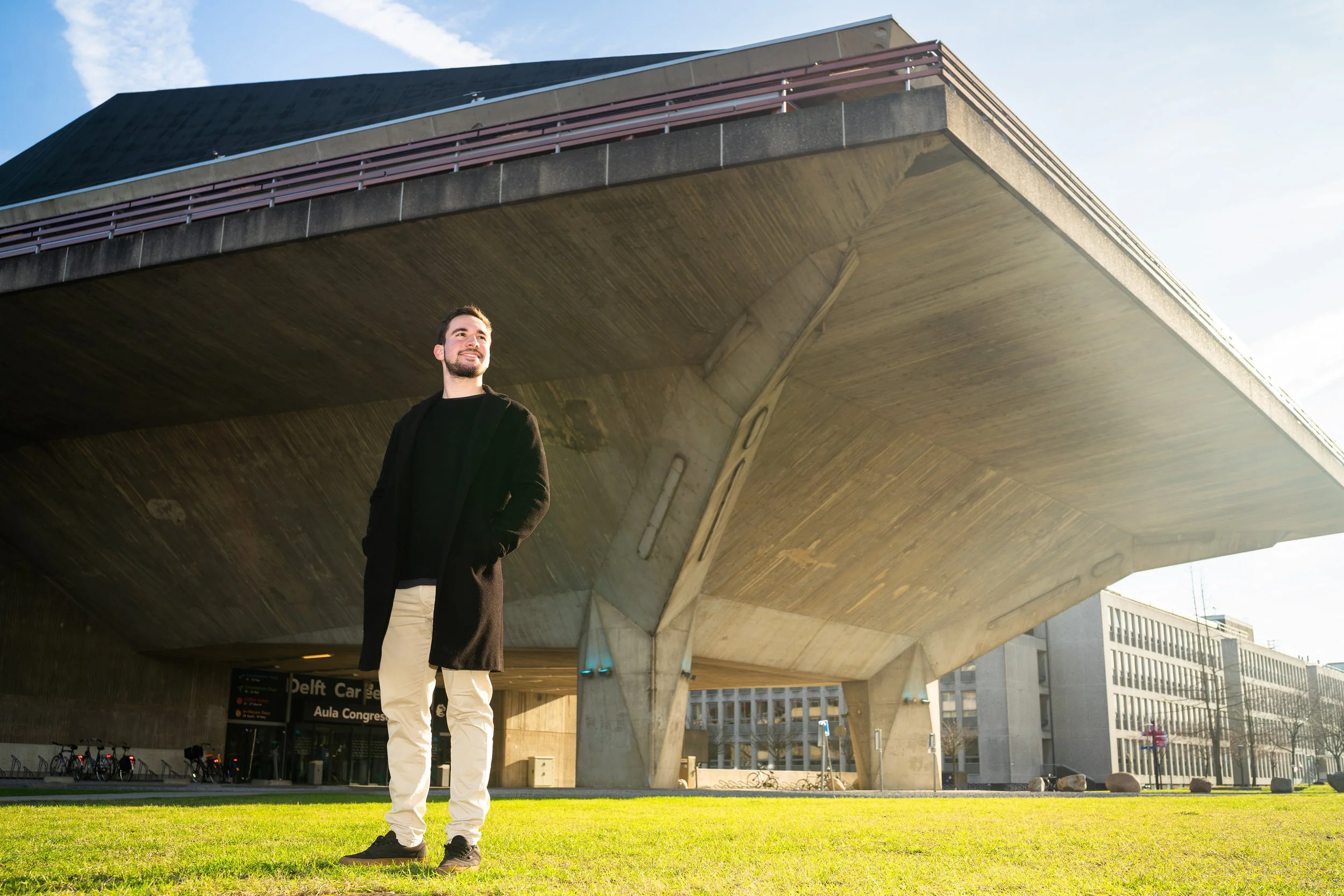 A man stands on green grass in front of a modern building with a large overhanging concrete roof, smiling and looking upward during daytime.