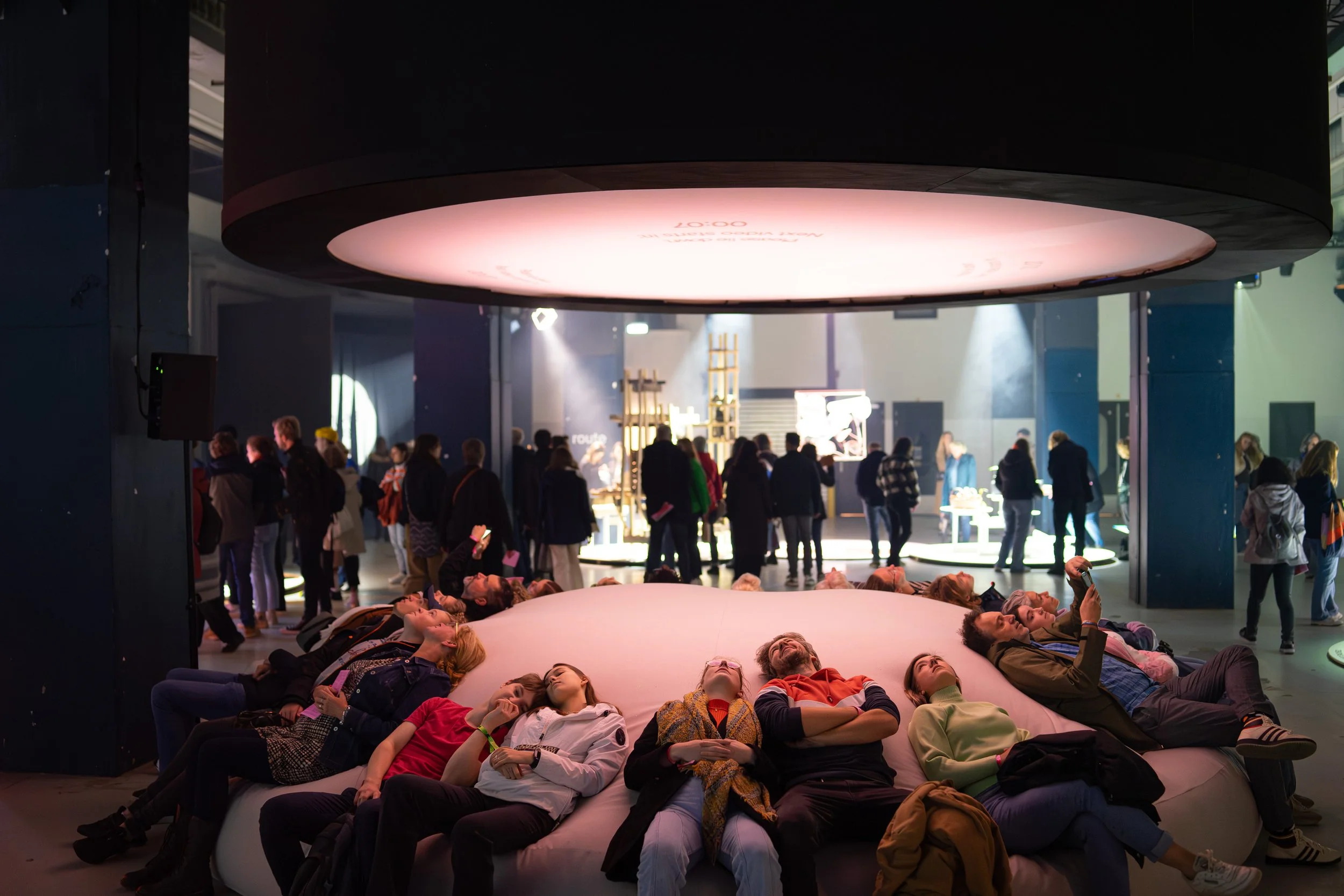People lying on a large circular cushion under an overhead display at an indoor event or exhibition.