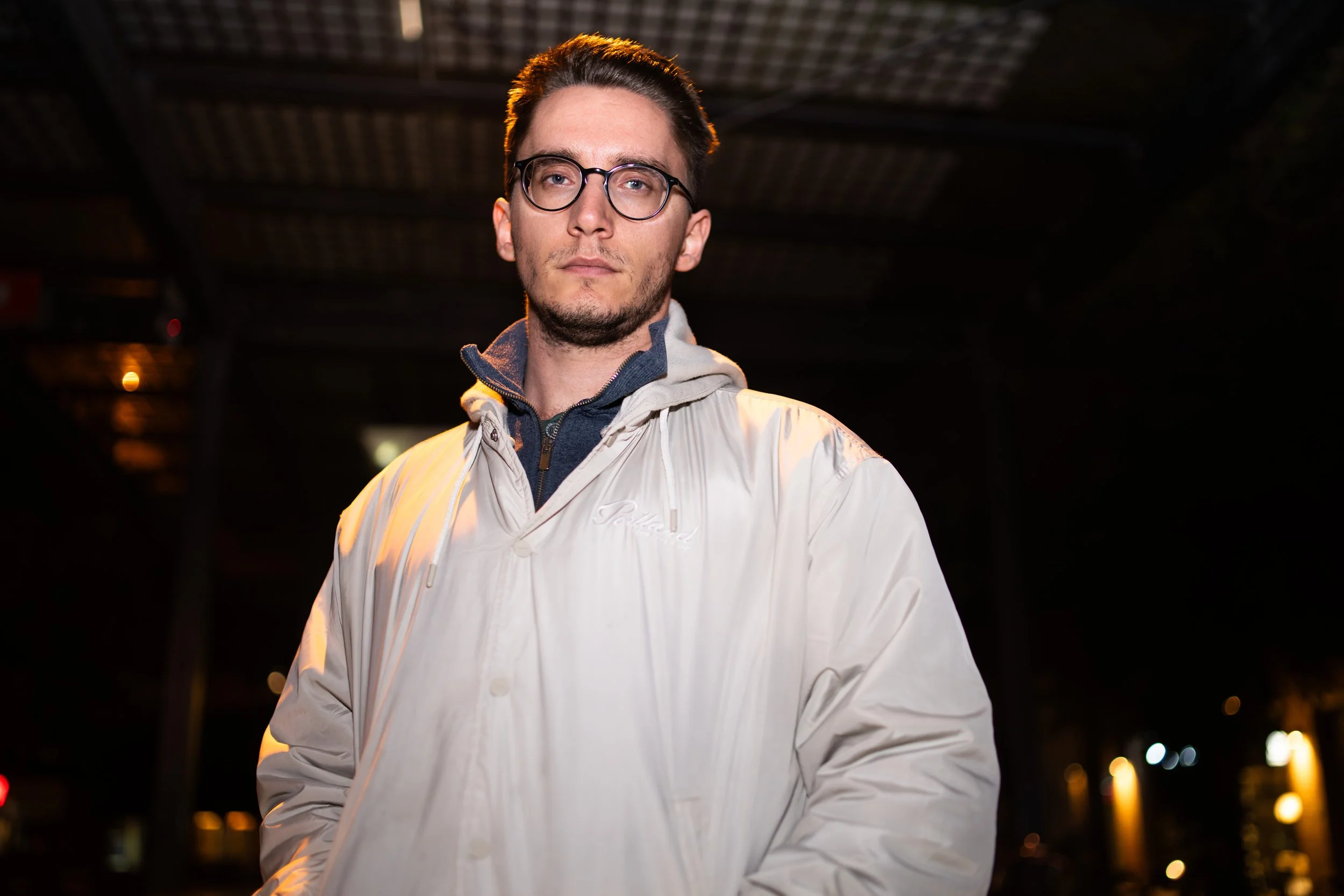 A young man with glasses, wearing a light-colored jacket over a dark hoodie, stands outdoors at night under a bridge or overpass, with some blurred city lights in the background.