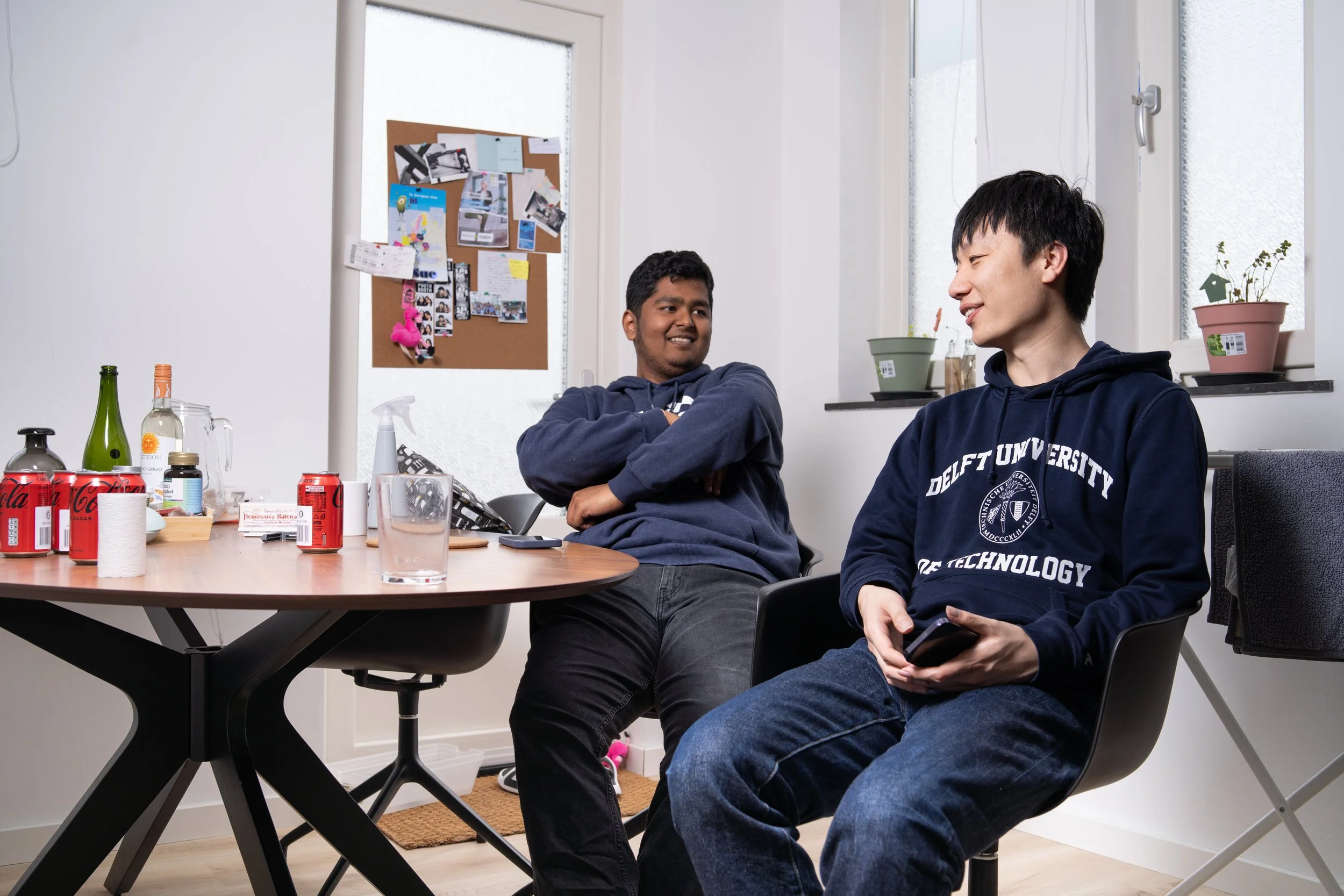 Two young men are sitting and talking in a modern room with a table, what appears to be an office space or a casual meeting area. One is smiling with arms crossed, and the other is holding a phone, both wearing hoodies.