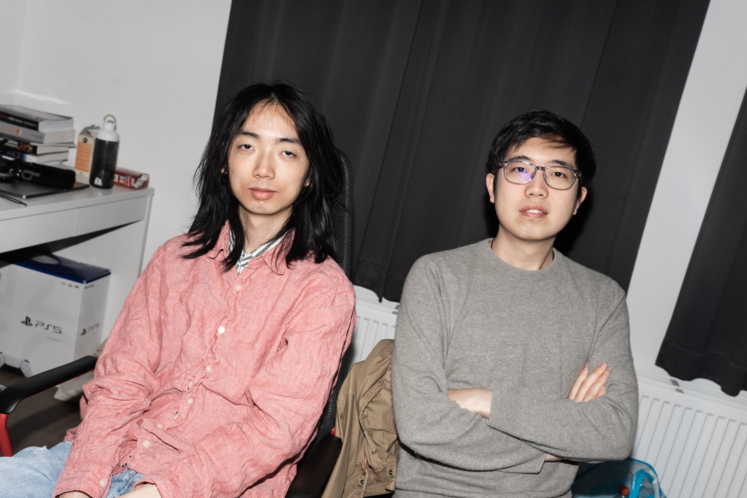 Two young men sitting in a room with white walls and dark curtains. One has long black hair, wearing a pink shirt and jeans, and the other has short black hair, glasses, and is wearing a gray sweater. A desk with books, a water bottle, and a PlayStat