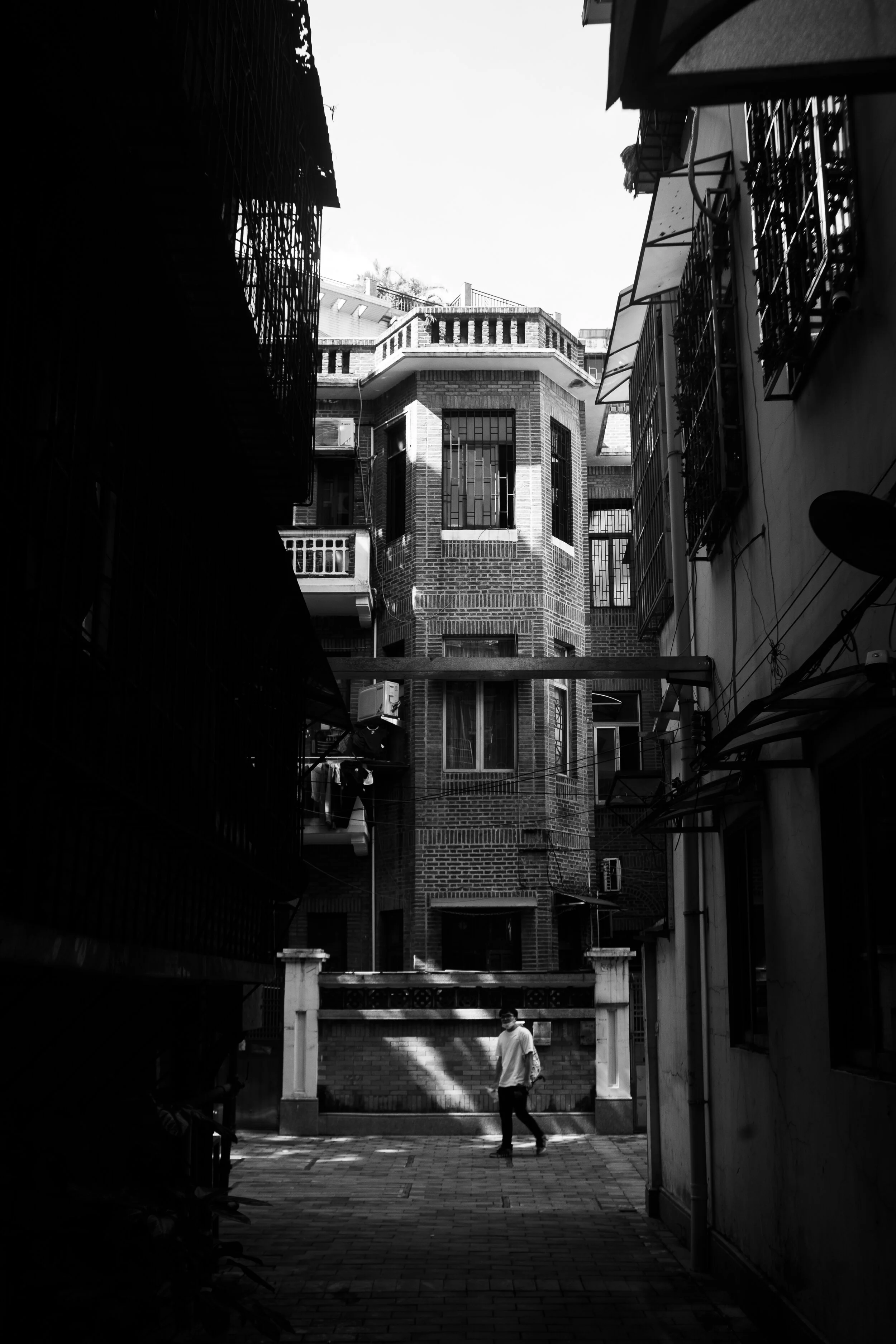 A black-and-white photo of a narrow urban alley with tall apartment buildings, a man walking across the alley, and bright sunlight casting shadows.