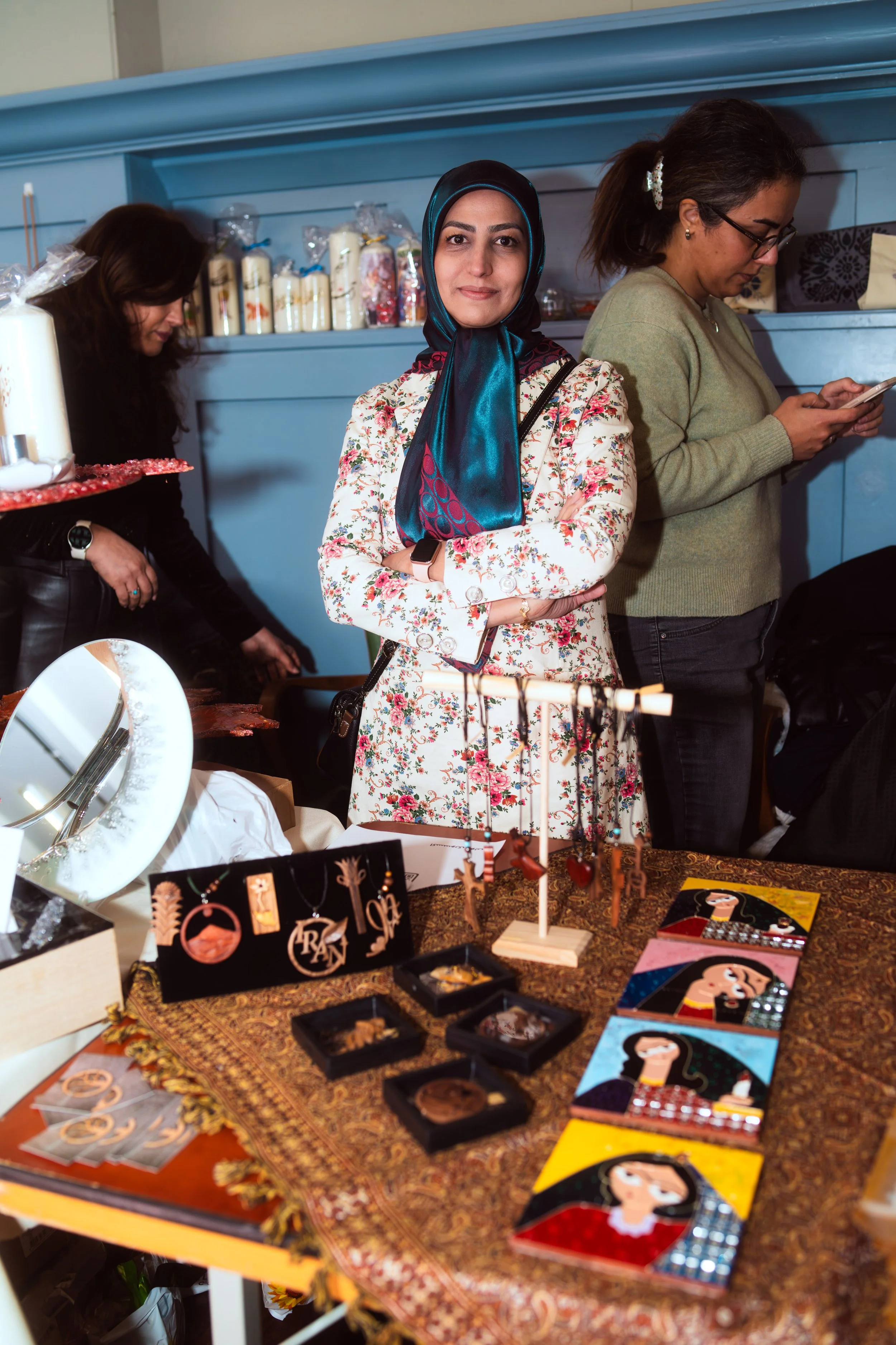 Woman wearing a floral dress and a hijab standing behind a jewelry display at an indoor market or craft fair.